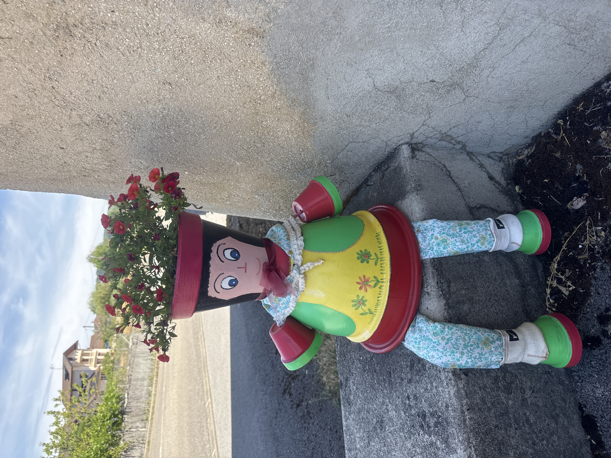 A decorative figure made from painted terracotta pots sits on a concrete ledge outdoors. The figure has a head shaped from a pot, painted with blue eyes, a pink nose, and black hair. Red flowers are planted in the top pot, serving as the figure's "hair." Its body is constructed from a larger pot painted with a yellow bodice adorned with small floral designs, a green "skirt," and a red base. Red terracotta pot saucers are attached to the sides of the body, painted green on the inside, serving as arms. The figure has legs made from fabric with a floral pattern, white socks, and green shoes with red soles. It is positioned on a grey concrete ledge next to a rough, light-colored stucco wall. The background shows a street, trees, and a building. The location is Coteaux du Lizon, France.