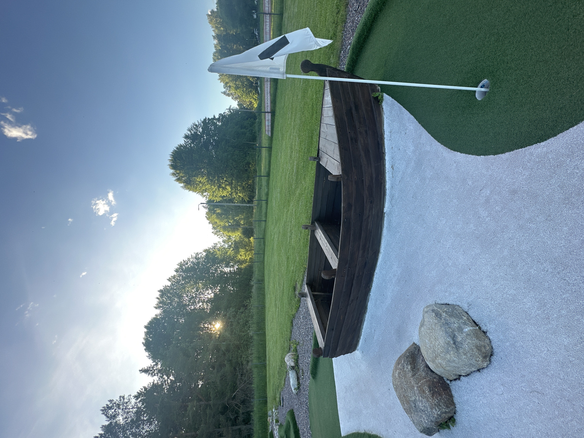 The image depicts an outdoor miniature golf course on a sunny day. In the foreground, a wooden boat structure is placed on a white gravel pathway. Two large rocks rest on the gravel near the boat. A white golf flag with a black marking is planted in a hole on the green artificial turf, to the right of the boat. The background features a lush green lawn, a line of trees, and a clear blue sky with scattered clouds. The sun is setting, casting a warm glow and long shadows. The location is an outdoor recreational area with a focus on mini golf.