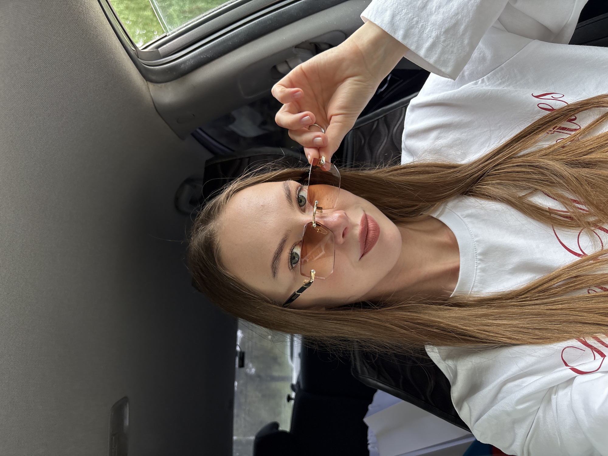 A young woman with long, light brown hair, wearing sunglasses and a white t-shirt with red writing, is sitting in the passenger seat of a car. She is holding the arm of her sunglasses with her right hand. The interior of the car is visible, including the dashboard, ceiling, and seats. Outside the car window, trees and foliage can be seen, suggesting an outdoor, possibly rural or suburban setting. The lighting indicates it is daytime. No specific landmarks or signs of the city of Perm, Russia are identifiable in the image.