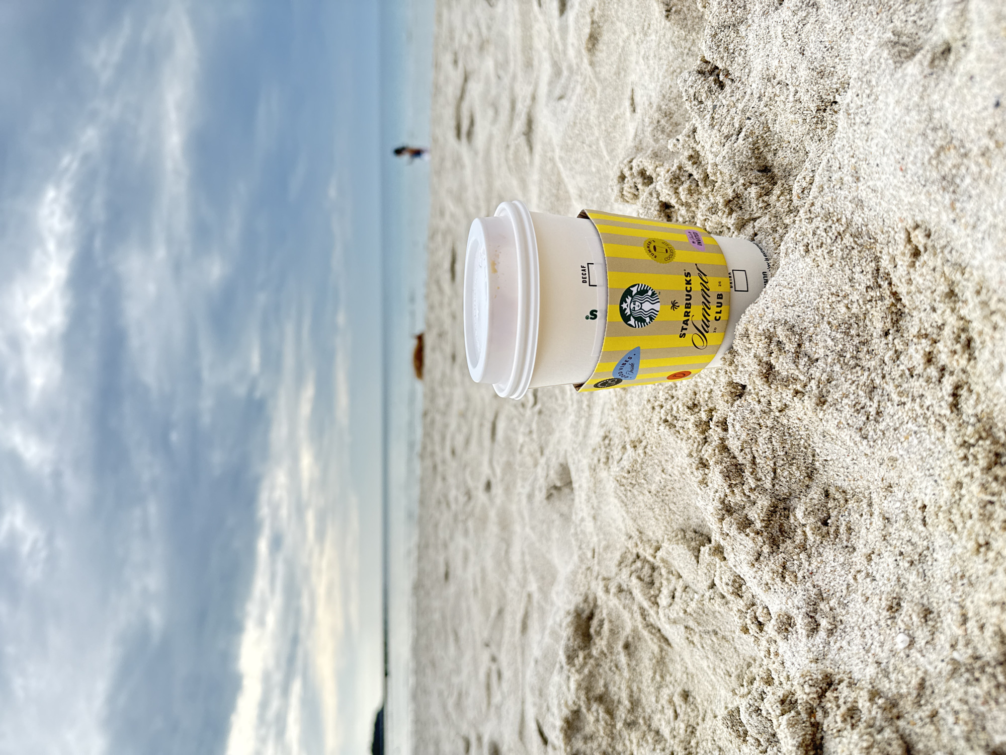 A Starbucks coffee cup with a yellow sleeve is placed on the sandy beach of Ko Samui District, Thailand. The cup is positioned in the foreground, with its lid on and a portion of the sleeve visible, featuring the Starbucks logo and "Summer Club" text. The background shows the ocean and a cloudy sky. Two people are faintly visible in the distance on the beach, along with a reddish-brown object lying on the sand. The sand is light in color and textured with small dunes.