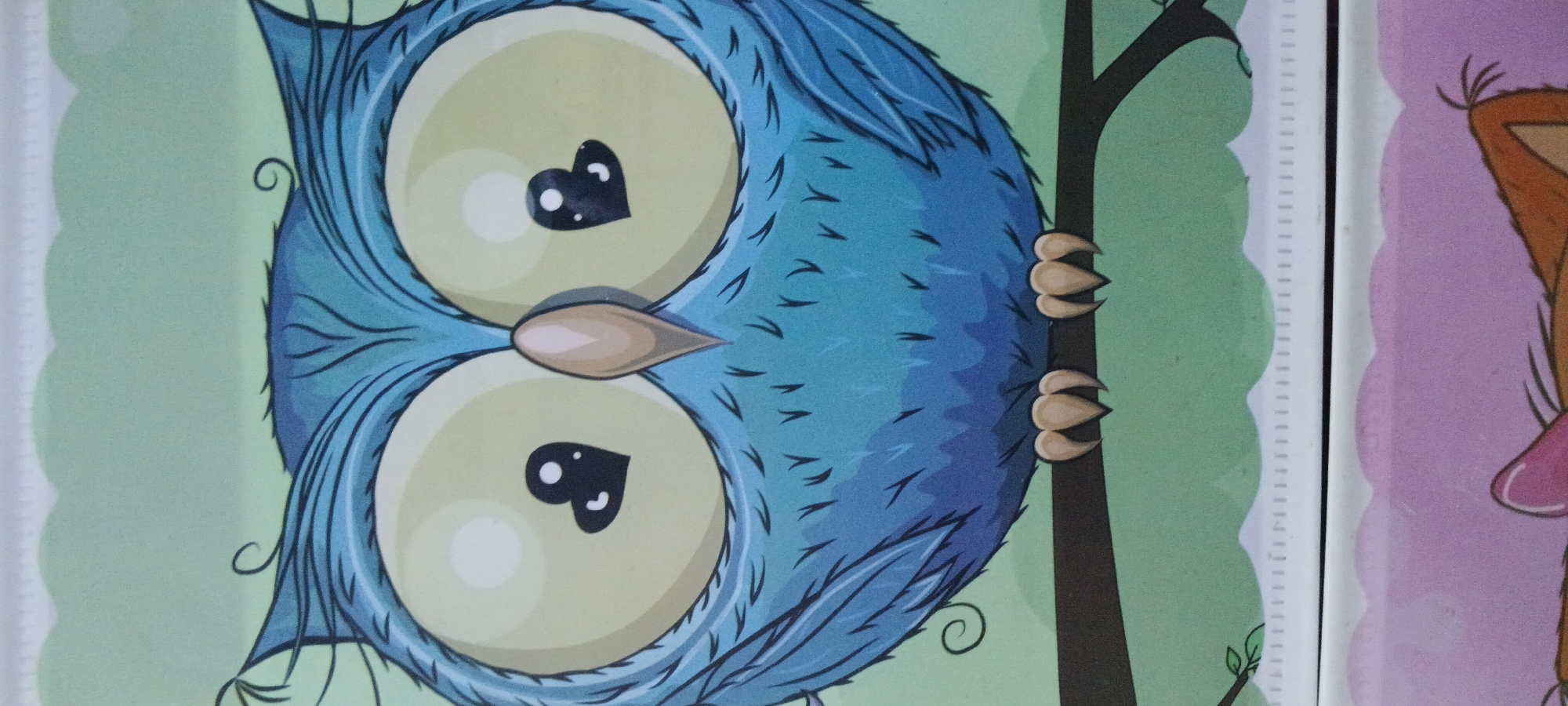 A close-up image features a stylized illustration of a cartoon owl. The owl is predominantly blue with large, light yellow eyes, each containing a small black heart shape with two white dots. Its beak is a light brown, and it has prominent feather details. The owl is perched on a dark brown branch against a light green background with a decorative scalloped edge at the top and bottom. Below the owl, the image transitions to a pinkish-purple background with a partial illustration of another cartoon character visible. The image appears to be a detail from a larger piece of artwork or product, possibly a decoration or stationery item. There is no discernible connection to Rejing, Indonesia, in the visual content of the image.