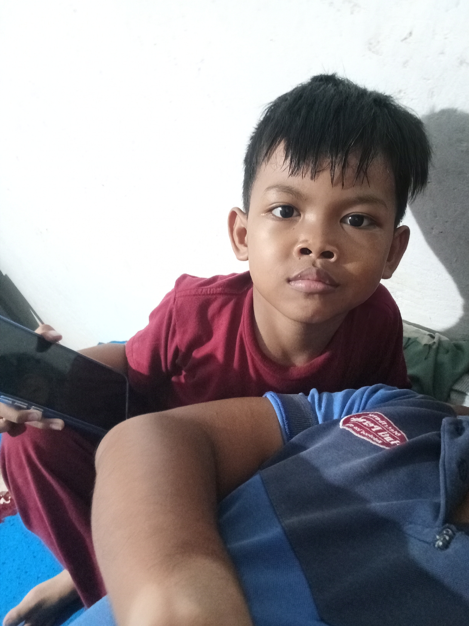 This is a close-up, eye-level selfie of a young boy in Cirebon, Indonesia. He is looking directly at the camera with a neutral expression. The boy has dark, short hair and is wearing a dark red t-shirt. He is holding a black tablet in his left hand. In the foreground, a person wearing a blue sweatshirt is partially visible, with their arm and shoulder in the frame. The background consists of a plain white wall with a shadow cast on it, suggesting indoor lighting. The overall impression is casual and domestic. There is no discernible text in the image.