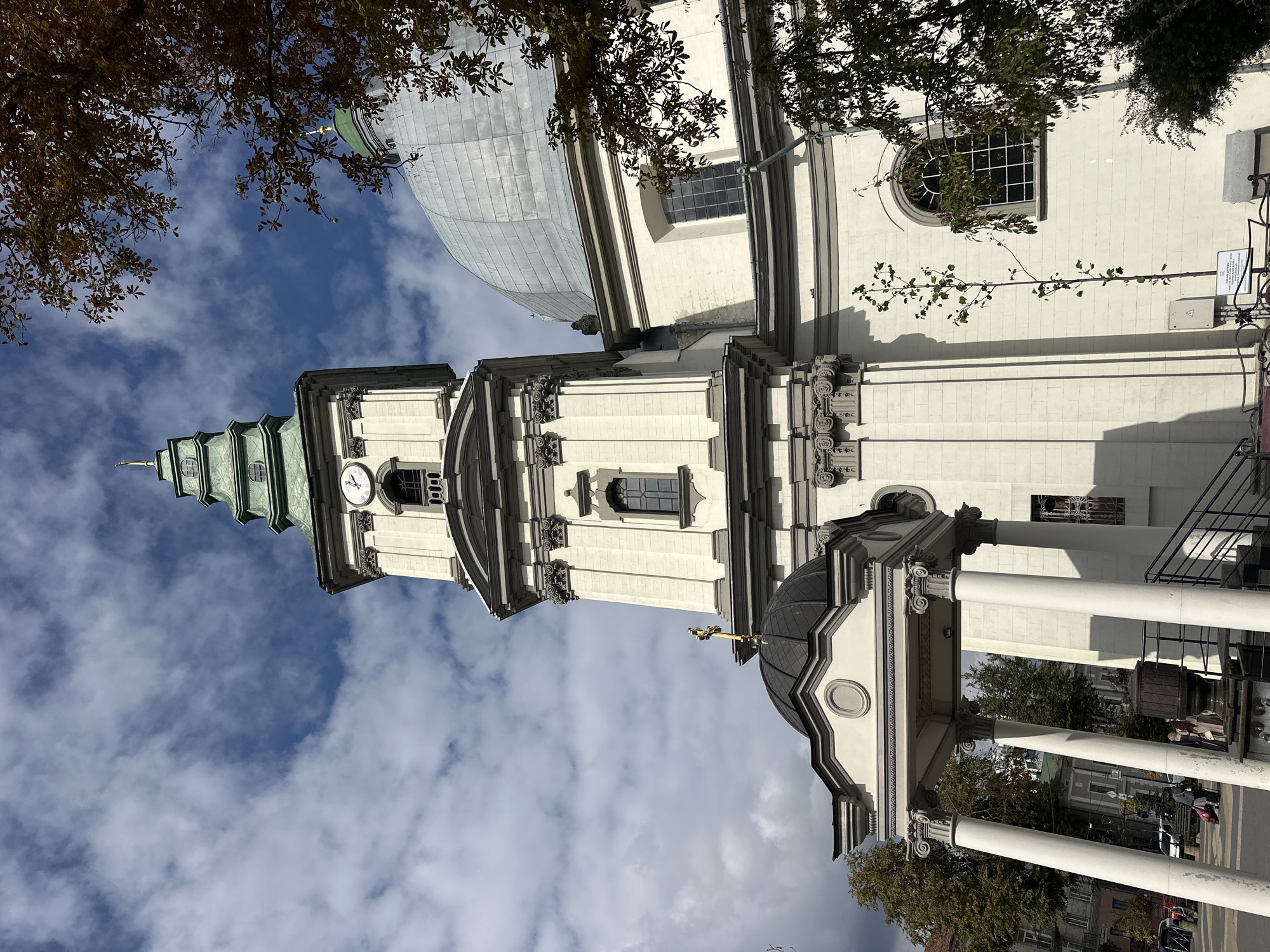 The image shows the Ternopil Cathedral, also known as the Cathedral of the Immaculate Conception of the Blessed Virgin Mary, in Ternopil, Ukraine. The cathedral is a large, white stone building with a prominent clock tower topped with a green spire. A large, silver dome is visible on the right side of the building. The sky is partly cloudy with blue patches. A tree with autumnal foliage is visible on the right side of the image, partially obscuring the dome. In the foreground, a portico supported by four white columns leads to a set of stairs. Some people are visible in the background, walking on a paved area in front of the cathedral. The architectural style appears to be Baroque or Neoclassical.