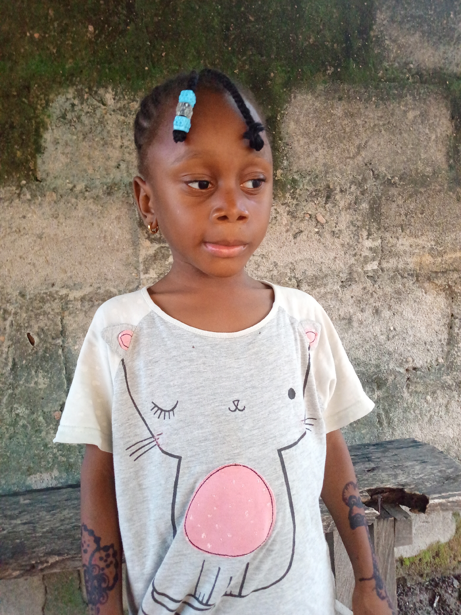 A young girl stands in front of a weathered concrete wall with moss growth. She has dark skin, small gold hoop earrings, and her hair is styled in braids with a blue and silver hair accessory. She wears a short-sleeved shirt featuring a cartoon cat design with a pink belly. There appear to be dark temporary tattoos on both of her forearms. To her right is a damaged wooden bench. The location is in the city of Unknown, Unknown.