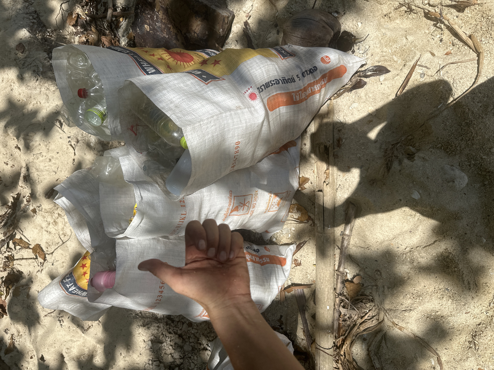 The image depicts a beach scene in ตำบลเกาะพะงัน, Thailand, with several white woven bags filled with plastic bottles and other debris. A hand is visible on the left, giving a thumbs-up gesture, suggesting a positive action related to the collected waste. The bags are placed on the sandy ground, which is scattered with dried leaves and twigs, indicating a natural environment. The lighting suggests it is daytime, with shadows cast by foliage and possibly the person taking the photo. One of the bags has visible text and graphics, including a sun-like emblem and what appears to be Thai writing. A coconut is also visible near one of the bags. The overall scene conveys a sense of environmental cleanup efforts on a beach.