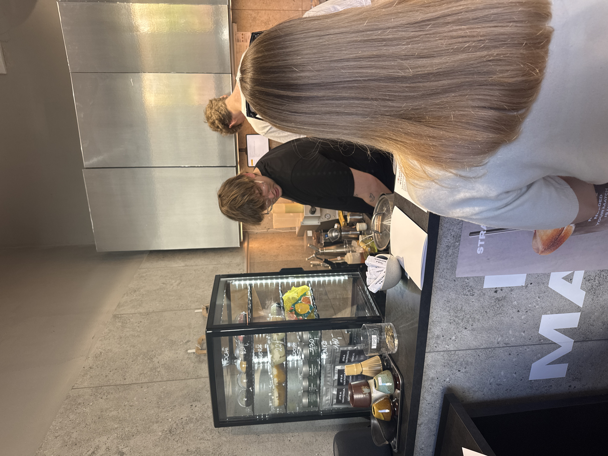 The image depicts a scene inside a café in Warsaw, Poland.
**People:** There are two individuals, an adult with blonde hair who is wearing a white shirt, and a second adult who appears to be a barista. The barista is wearing a black shirt and glasses.
**Objects:** A refrigerated display case sits near a countertop, filled with items such as mochi and matcha products. On the counter, there are matcha whisks, cups, and papers with the words "MATCHA" and "STRA" printed on them.
**Environment:** The café features a modern aesthetic, with a concrete floor and wall. Stainless steel surfaces are visible behind the counter.