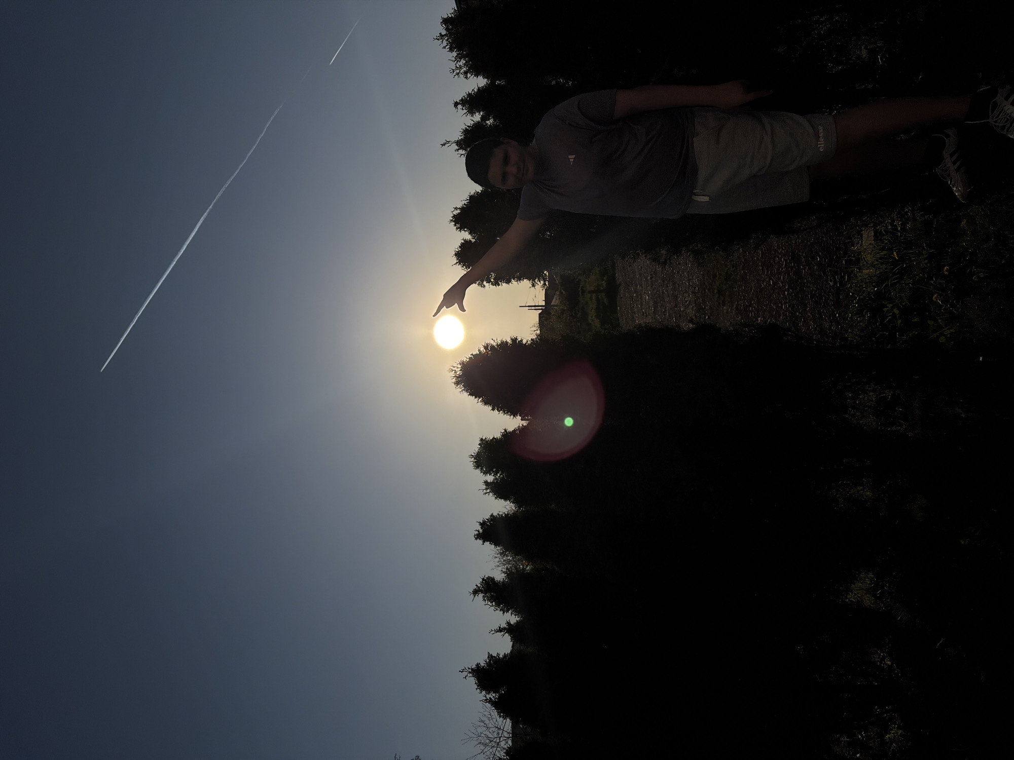 A person stands outdoors, pointing at the sun. The person is wearing a t-shirt, shorts and sneakers. A contrail is visible in the sky. Tall trees are in the background. The location is not identifiable as being in Sofia, Bulgaria.