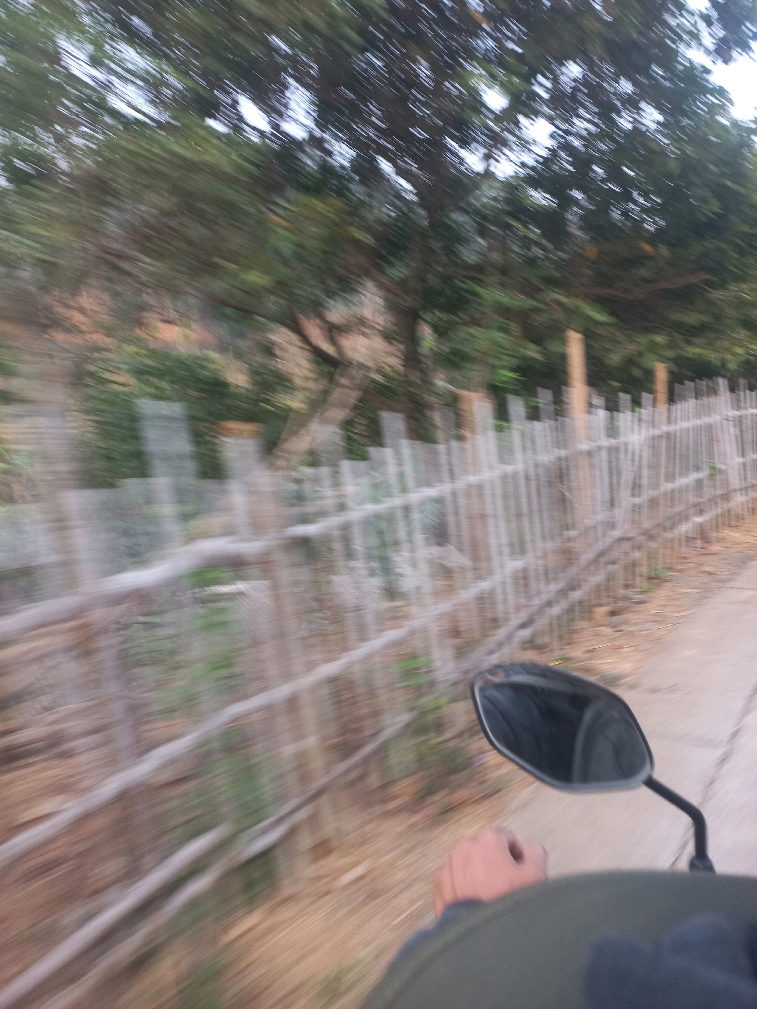 This is a first-person perspective photo taken from a moving vehicle, likely a motorcycle, in Sukoharjo, Indonesia. The image is motion blurred, indicating the vehicle's speed. In the foreground, a hand and a portion of the vehicle's body are visible, with a side-view mirror on the right. The immediate background features a dusty road next to a wooden fence, constructed from vertically placed poles and horizontal wooden slats. Beyond the fence, a blurred landscape of trees and vegetation is visible against a bright sky. The overall scene depicts a rural or roadside environment during daytime.