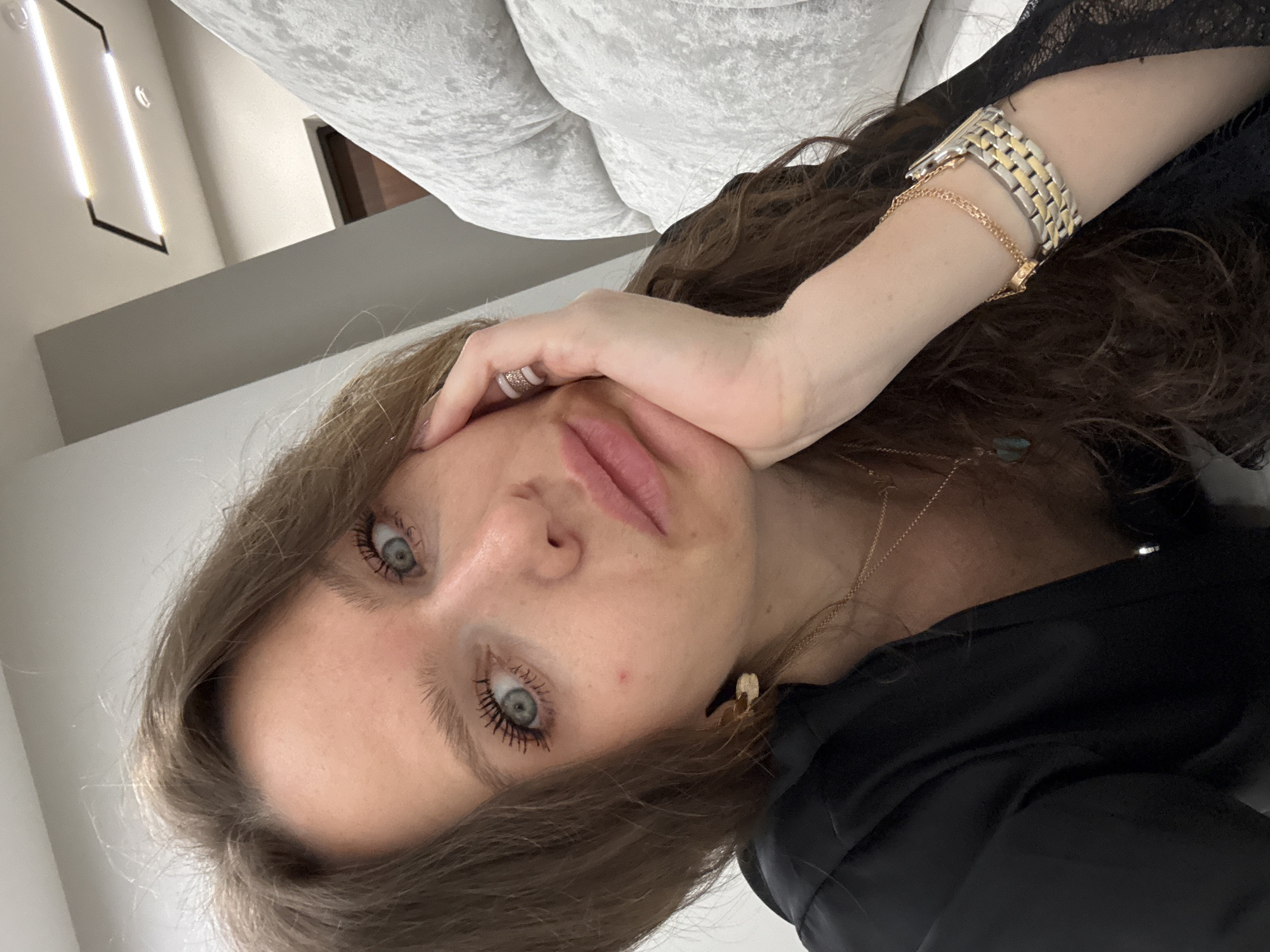 A selfie of a woman with long brown hair and blue eyes is captured in an indoor setting. She is positioned close to the camera, partially propped up by her hand, which is adorned with a ring. She is wearing a watch and bracelet. A white couch is visible in the background, alongside a modern lighting fixture on the wall. The woman is wearing a black garment. The exact location is undetermined.