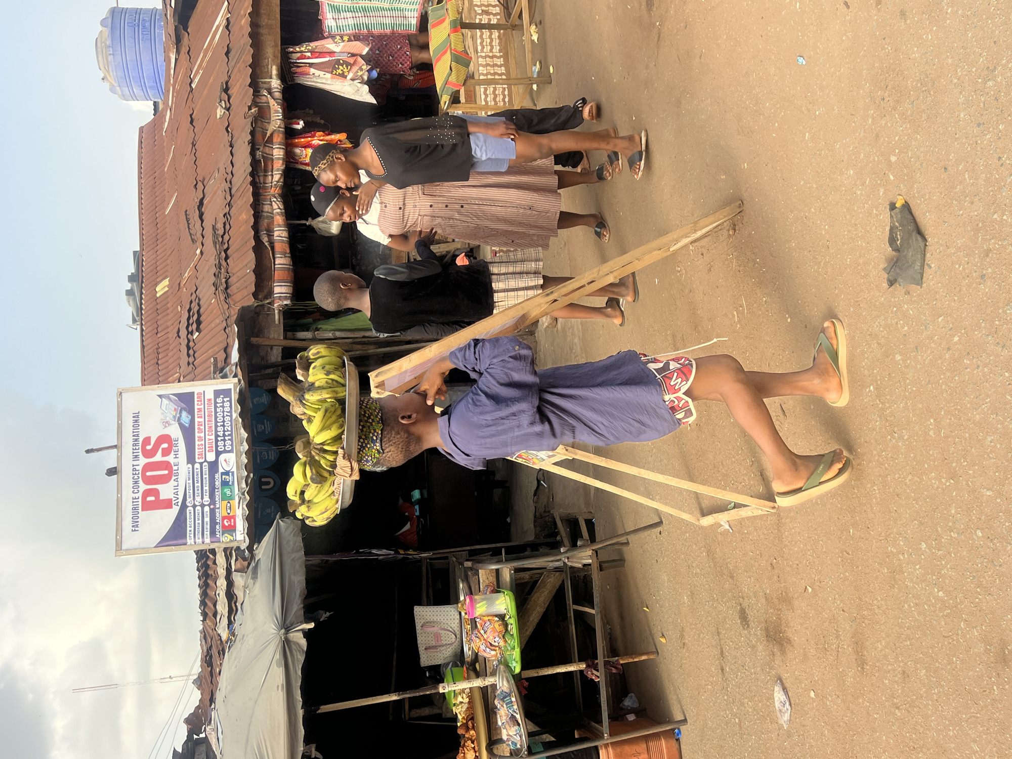 The image captures a vibrant outdoor market scene, likely in Obosi, Nigeria, bathed in bright daylight under a clear sky. In the foreground, a young individual, dressed in a blue shirt and patterned shorts, skillfully balances a large, round tray laden with yellow bananas on their head. They walk forward, also holding a light wooden frame, possibly for a stall or carrying another load.
Behind this central figure, several other people are present. A group of three women or girls stands near market stalls: one in a black top and blue shorts, another in a striped dress, and a third partially visible in a dark top. Another person in a dark shirt stands between the banana vendor and this group.
The market features numerous stalls constructed with corrugated iron roofs, displaying various goods such as colorful fabrics. A large blue water tank is visible on a distant rooftop. The ground is a paved, somewhat uneven surface, with occasional debris like plastic bags.
A prominent white sign in the upper left, belonging to "FAVOURITE CONCEPT INTERNATIONAL," advertises "POS AVAILABLE HERE." The sign lists services including "OPEN ACCOUNT, DEPOSIT MONEY, WITHDRAW MONEY, SEND MONEY, BUY AIRTIME DATA, PAY YOUR BILLS," along with "SALES OF OPAY ATM CARD" and "DAILY CONTRIBUTION." Contact numbers "08148100516" and "09112097881" are displayed, and the location is specified as "AFOR-ADIKE MARKET OBOSI." The scene depicts typical daily commerce and activity in a local market environment.