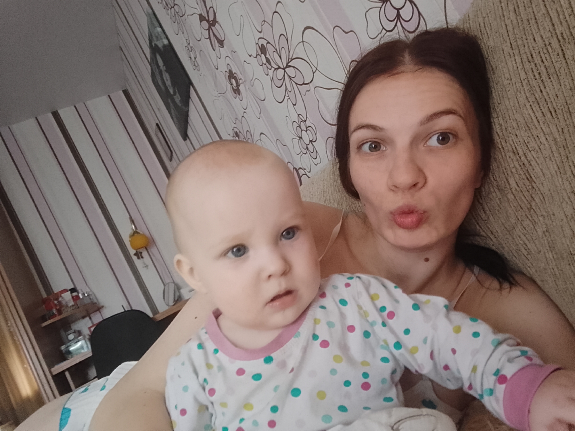 A woman and a baby are lying on a sofa in a room. The woman has brown hair pulled back into a bun and is making a kissing face. She is wearing a light-colored bra. The baby has a bald head and is wearing a white pajama top with multi-colored polka dots. The background shows floral wallpaper and striped wallpaper. A shelf with various items is visible to the left. The scene appears to be indoors, likely a residential setting.