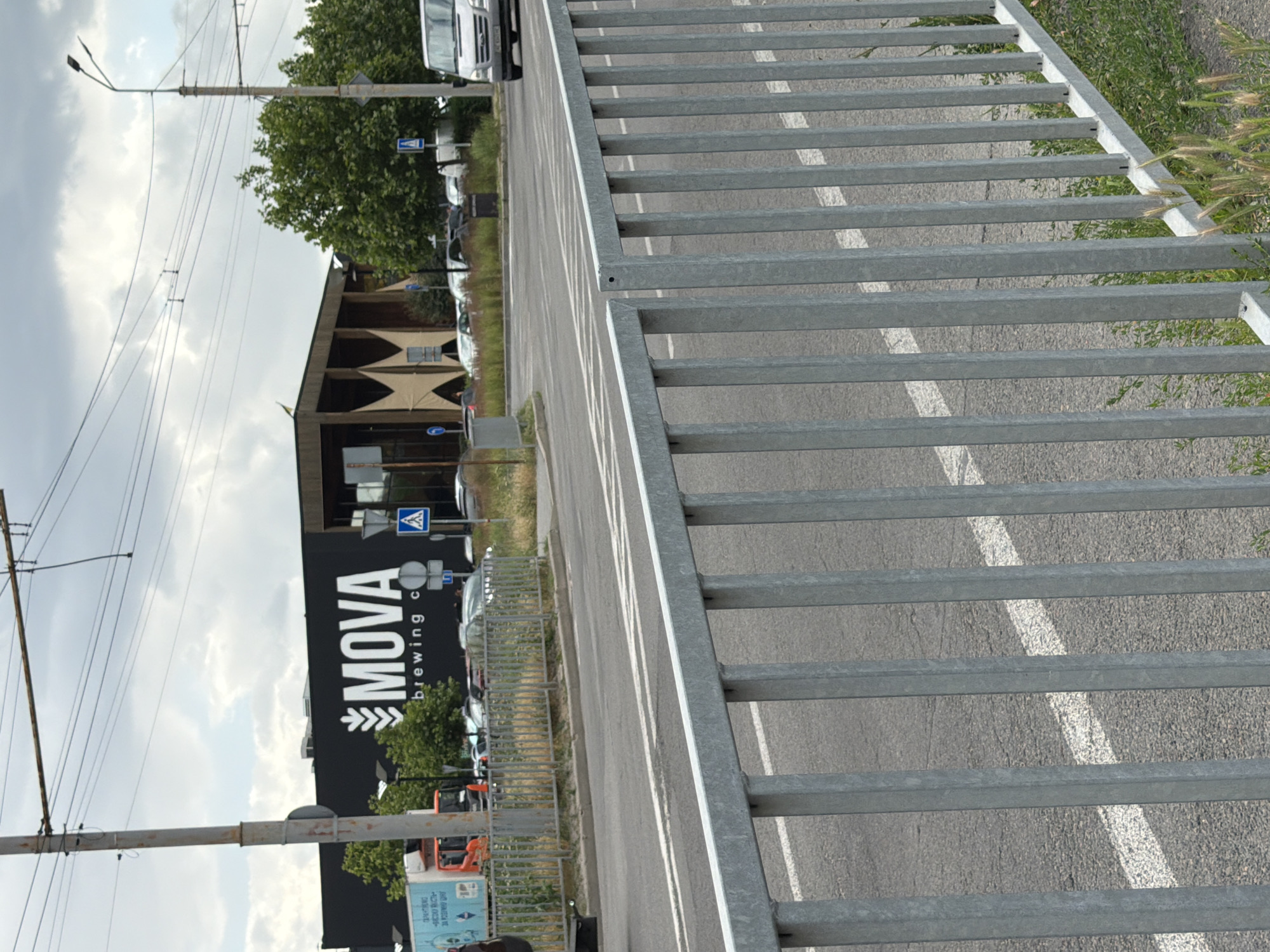 The image shows a street scene in Dnipro, Ukraine, with a focus on a building with a prominent "MOVA brewing co." sign. In the foreground, a metal fence runs horizontally across the frame, obscuring part of the street and sidewalk. The street itself appears to be a multi-lane road with white markings. On the left side of the image, a blue and white truck is parked. Behind the fence, a modern building with dark siding and angular architectural elements is visible. The "MOVA brewing co." logo is displayed in large white letters on the dark facade. Several trees are interspersed around the building. On the right side of the image, a white van is driving on the street. The sky is overcast with clouds. The environment appears to be an urban area, likely a commercial or industrial zone, given the presence of the brewery and the truck.