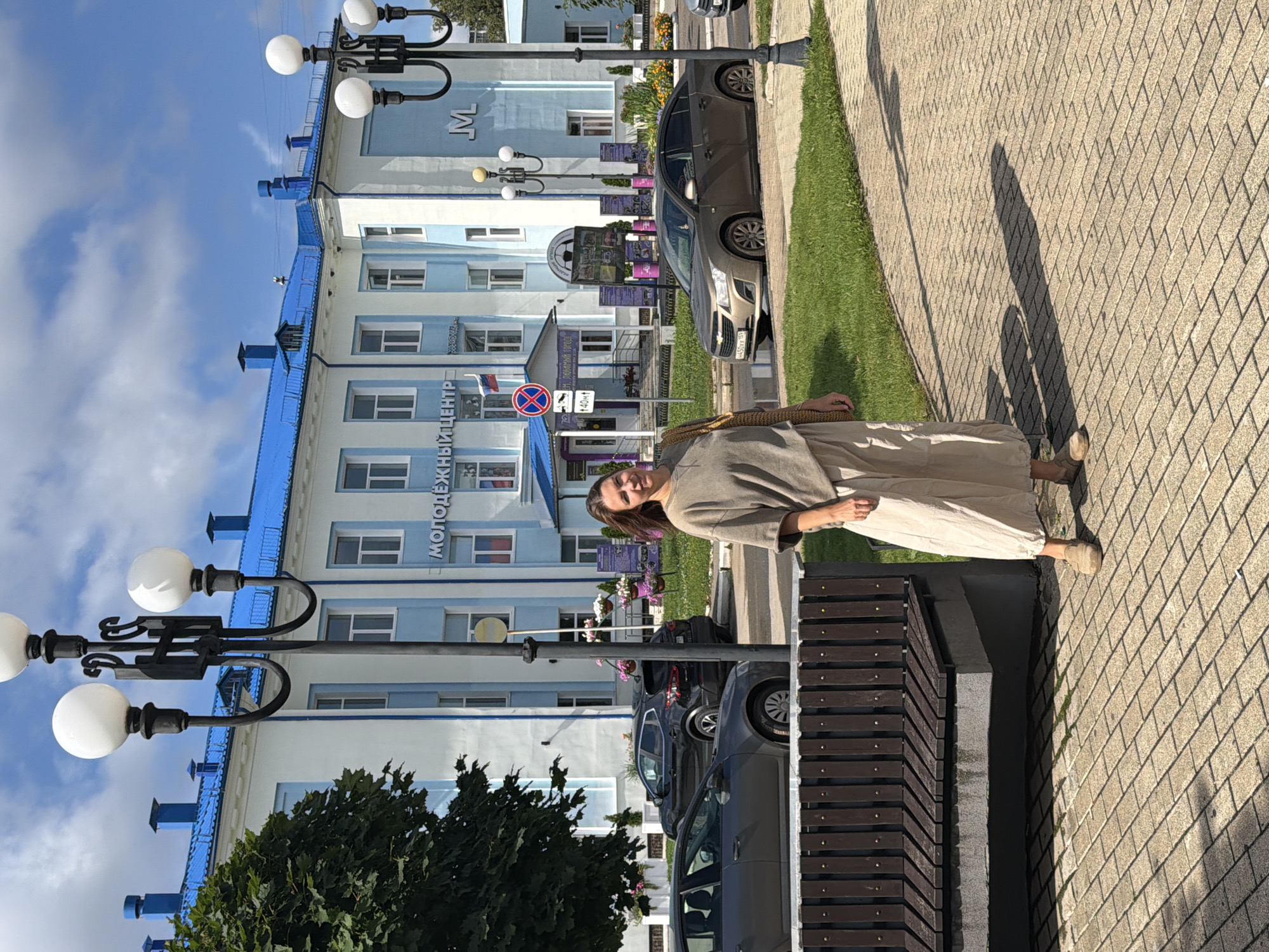 A woman stands on a paved sidewalk in front of a light blue and white building with a blue roof. The building has large windows and a sign that reads "МОЛОДЁЖНЫЙ ЦЕНТР" (Youth Center). Streetlights with multiple globes stand on the sidewalk. A bench is visible to the left of the woman, and several cars are parked on the street behind her. The woman is wearing a light-colored t-shirt and a long, flowing skirt. She has long, dark hair and is smiling at the camera. The scene appears to be set in the city of Chaikovsky, Russia.