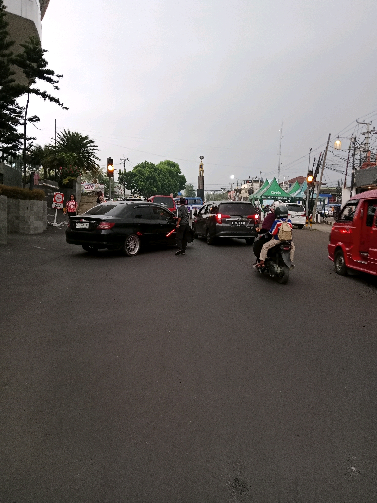In Cianjur, Indonesia, on an overcast day, traffic flows along a paved road. A black sedan, identified by its license plate "F 1308 XV," is stopped, with a person in a dark uniform and a reflective vest directing traffic with a glowing red baton. Behind the sedan, a dark SUV waits. On the right, a red commercial vehicle is visible. Further down the road, a motorcycle with two occupants, one wearing a backpack, travels away from the viewer. Traffic lights are visible, with one illuminated yellow. In the background, tents with the "Grab" logo are set up, suggesting a promotional event. Utility poles with numerous wires line the street, and a tall monument with a circular top stands in the distance. Trees and a building with a modern architectural style are present on the left side of the image.