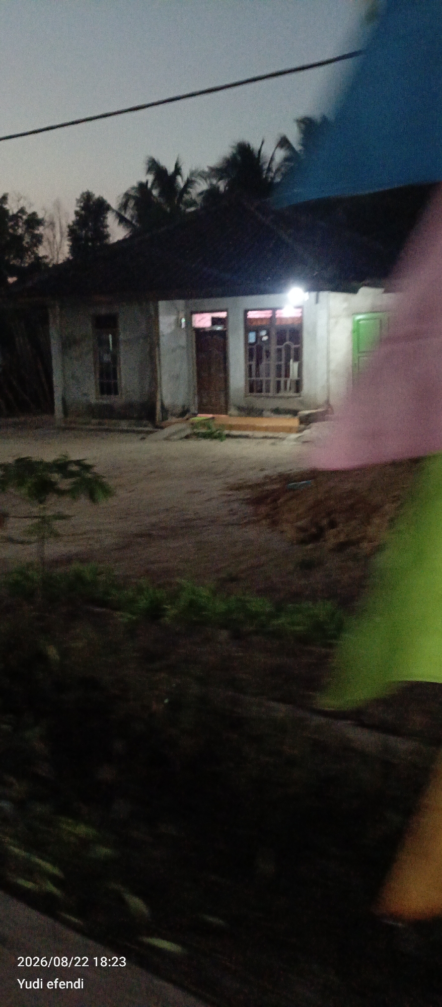 The image shows a small, single-story house with a dark roof, illuminated by a single light bulb above the entrance. The walls appear to be made of concrete or stucco. There are several windows and doors visible, some with glass panes and others with wooden doors. A green door is visible on the right side of the house. The area in front of the house is dirt or sand, with some sparse vegetation. Trees, including palm trees, are visible in the background against a twilight sky. A thin black wire runs across the upper portion of the image. The lower right corner of the image shows a blurred area of color, possibly from a flag or fabric, in pink and green. The bottom left corner displays text indicating "2026/08/22 18:23 Yudi efendi". The setting appears to be a rural area in Rawajitu Selatan, Indonesia.