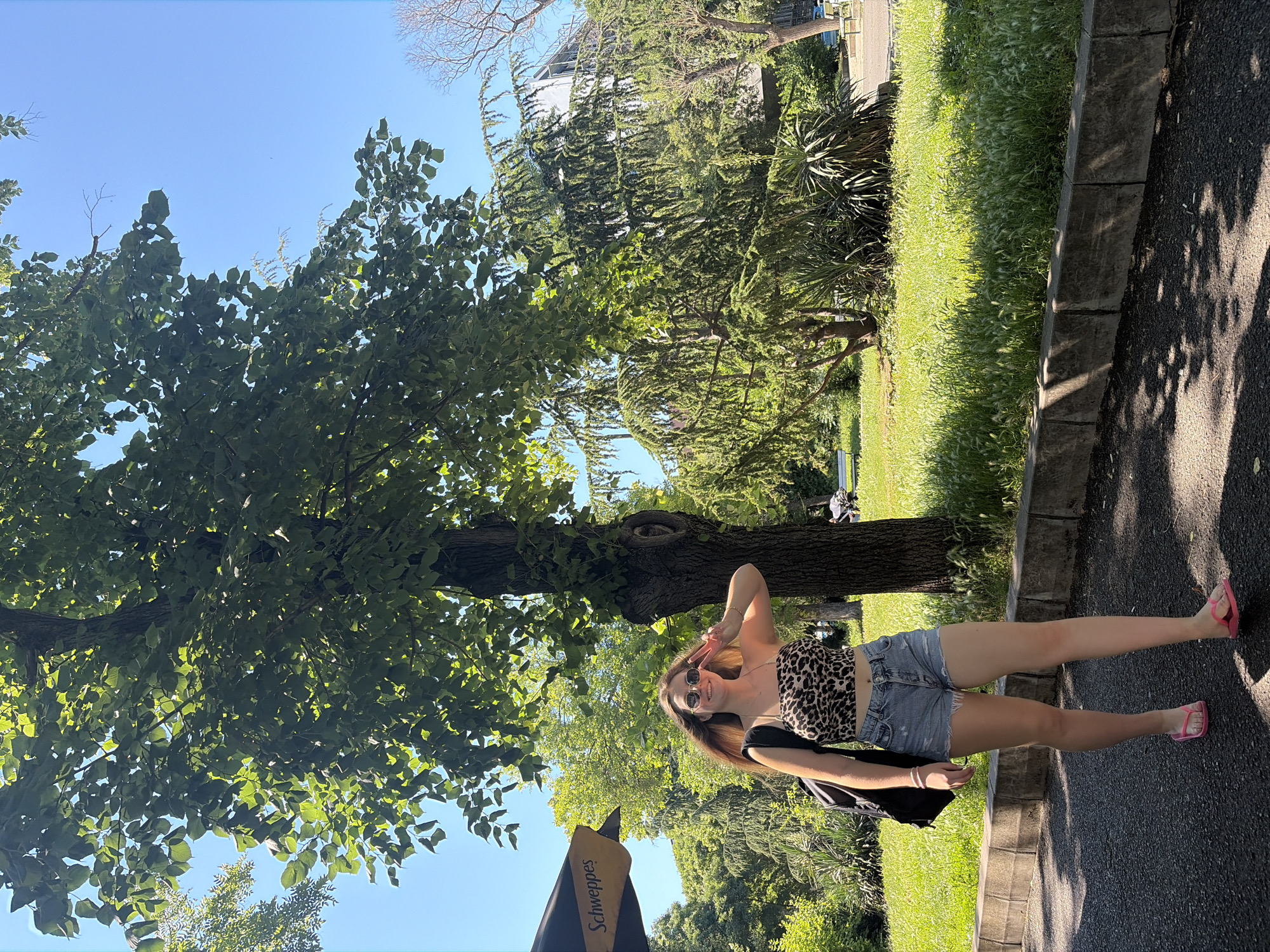 A young woman with long, blonde hair, wearing a leopard-print crop top, denim shorts, pink flip-flops, and sunglasses, is walking on a paved path in a park. She is smiling and holding up two fingers in a peace sign. A black backpack is slung over her left shoulder. She is located in the Sea Garden park in Burgas, Bulgaria, on a sunny day with a clear blue sky. The background features a large tree, lush green grass, and other trees and vegetation. A Schweppes umbrella is visible to the left of the frame.