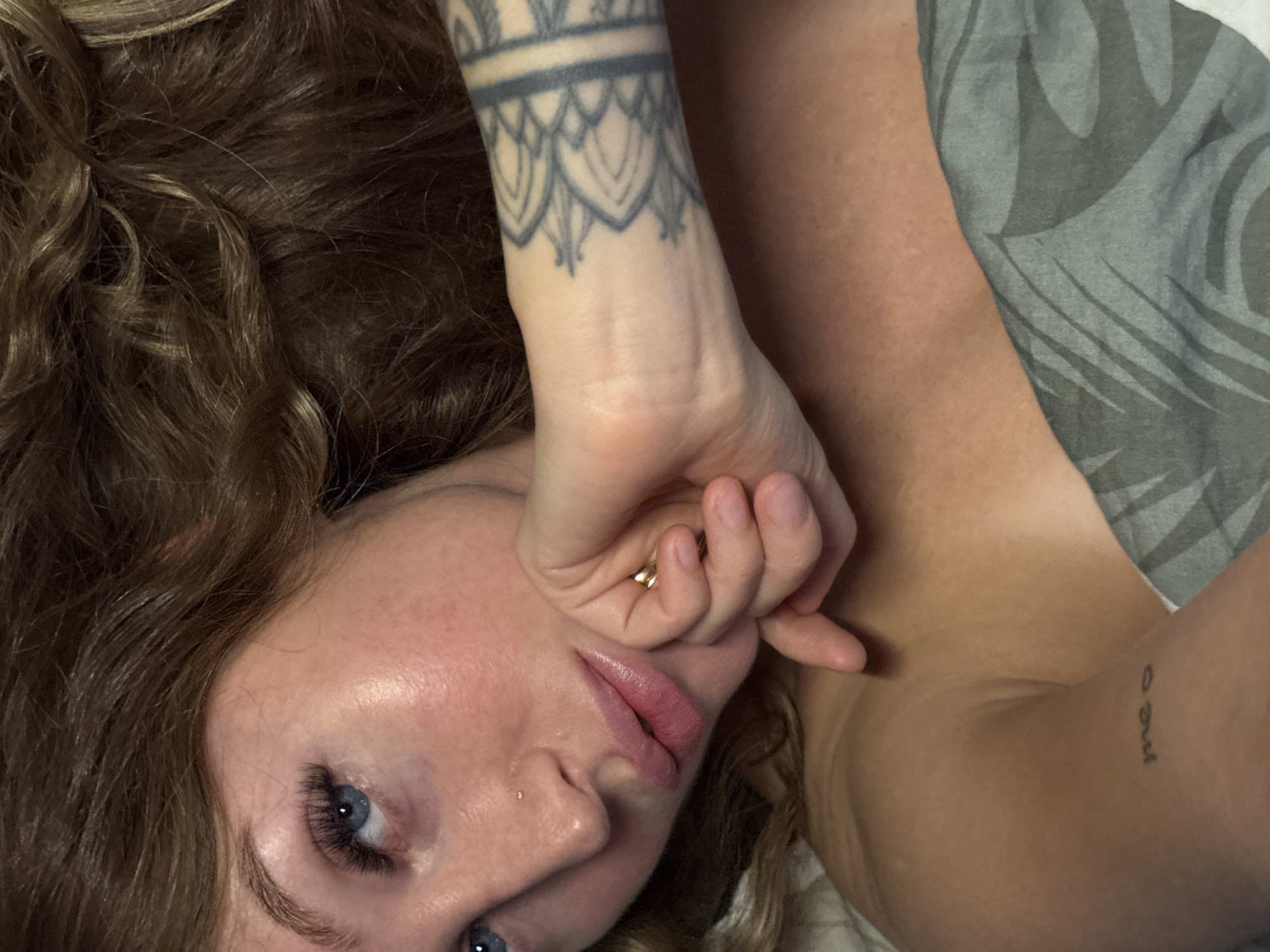 A person is lying down, facing the camera, with their hand near their face. They have curly brown hair, blue eyes, and fair skin. There's a tattoo on their wrist and another one visible on their upper arm. They are wearing what appears to be a grey tank top. The location is not discernible from the image, and there is no indication that it is in Chaikovsky, Russia.