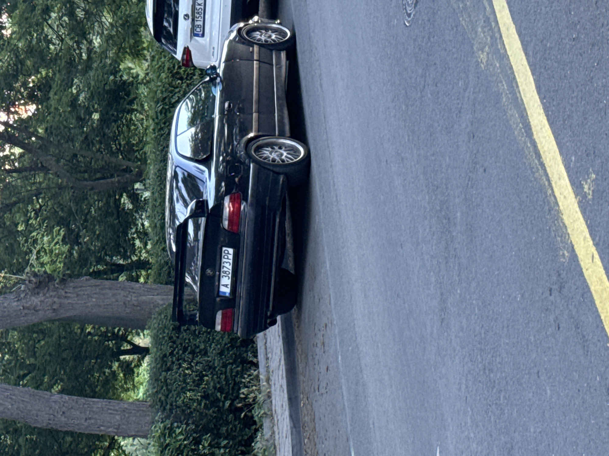 A black BMW E36 coupe with a large rear spoiler is parked on the right side of a paved street. The car's license plate reads "A 3873 PP". To the right of the BMW, a white SUV, possibly a Lexus, is also parked, with its license plate partially visible as "CB 1585 K". The vehicles are parked along a sidewalk lined with green bushes and mature trees, creating a leafy backdrop. The street has a yellow lane marking. The scene appears to be in Burgas, Bulgaria, based on the license plate format.
