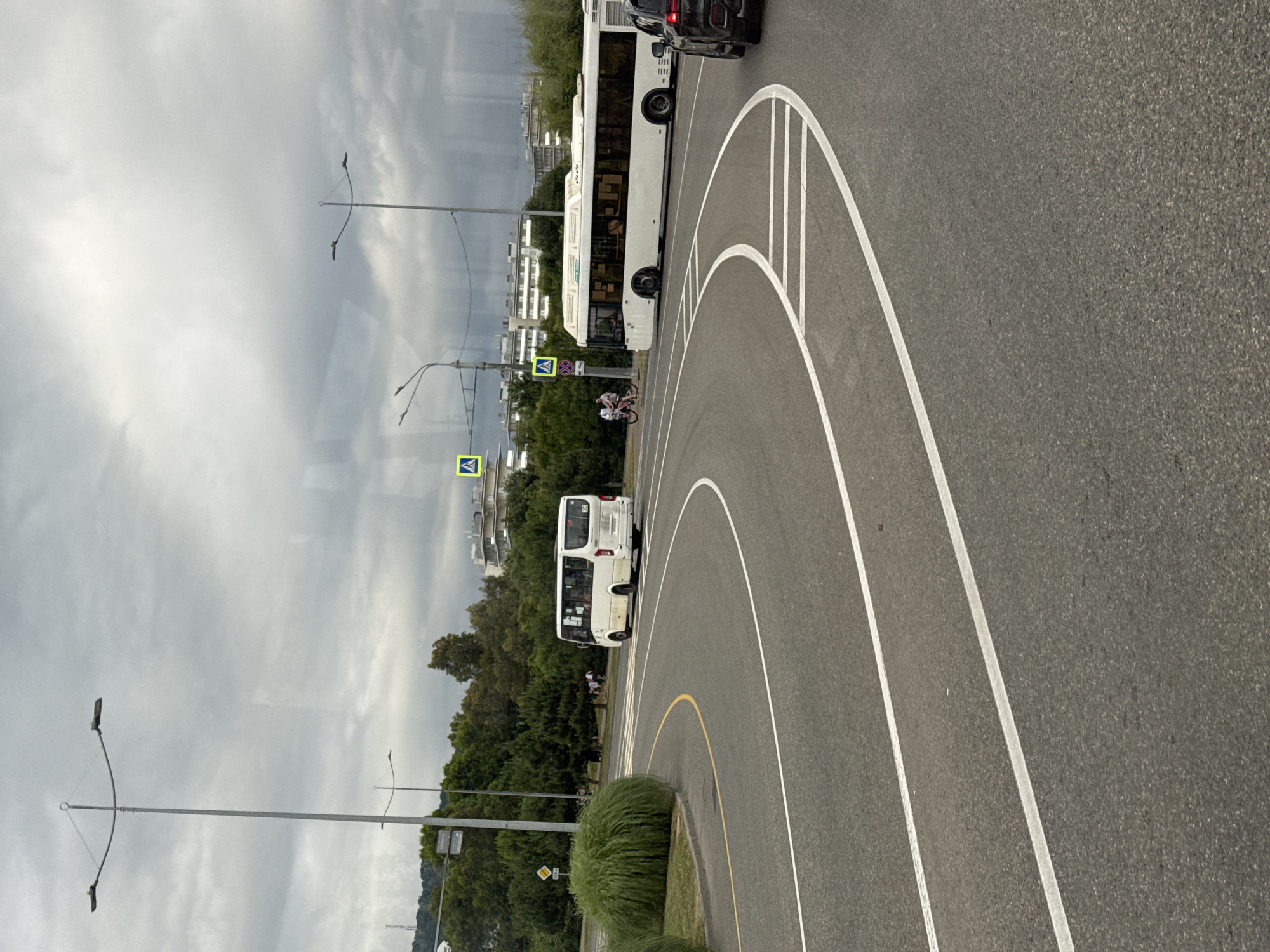 The media file depicts a street scene in Sochi, Russia, under an overcast sky. The primary subjects are two white buses, one larger and one smaller, on a paved road with clear lane markings. The road curves to the right, with white lines delineating the lanes and a yellow line marking the edge of the road or a turn. In the middle ground, a smaller white bus is visible, with people inside. To the left of the bus, a person on a bicycle is also present, along with several pedestrians. A pedestrian crossing sign is visible near the bicyclist. Further in the background, there are trees and buildings. Two streetlights are also visible in the scene, one on the left and one in the distance. The overall atmosphere is calm, with no strong indicators of a specific time of day other than it appears to be daytime. The weather seems to be cloudy, as suggested by the uniformly gray sky. No discernible text is visible in the image.