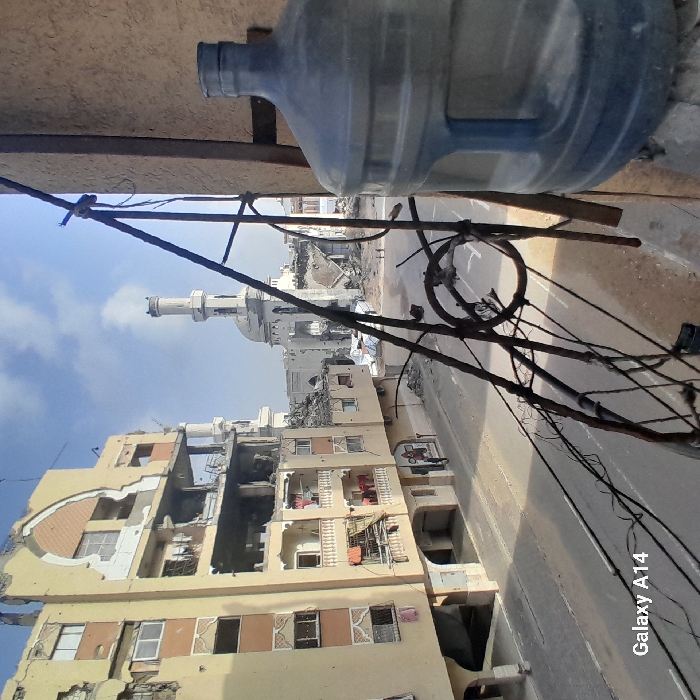 The image depicts a war-damaged urban street scene in Khan Yunis, Palestinian Territory, under a partly cloudy sky. In the immediate foreground on the right, a large blue plastic water bottle sits on a ledge, partially obscured by blurred, tangled, rusty metal wires or rebar extending diagonally into the frame. To the left, a multi-story, light yellow/beige building is severely damaged, with multiple floors exposed, sections of walls crumbled, and visible interiors; laundry hangs from several of its balconies. A paved street, largely empty save for some debris, runs across the midground. Two small, indistinct human figures are visible walking on this street in the central distance. In the background, a large mosque with a prominent light-colored minaret and a central dome stands heavily damaged, surrounded by extensive rubble and ruined structures.