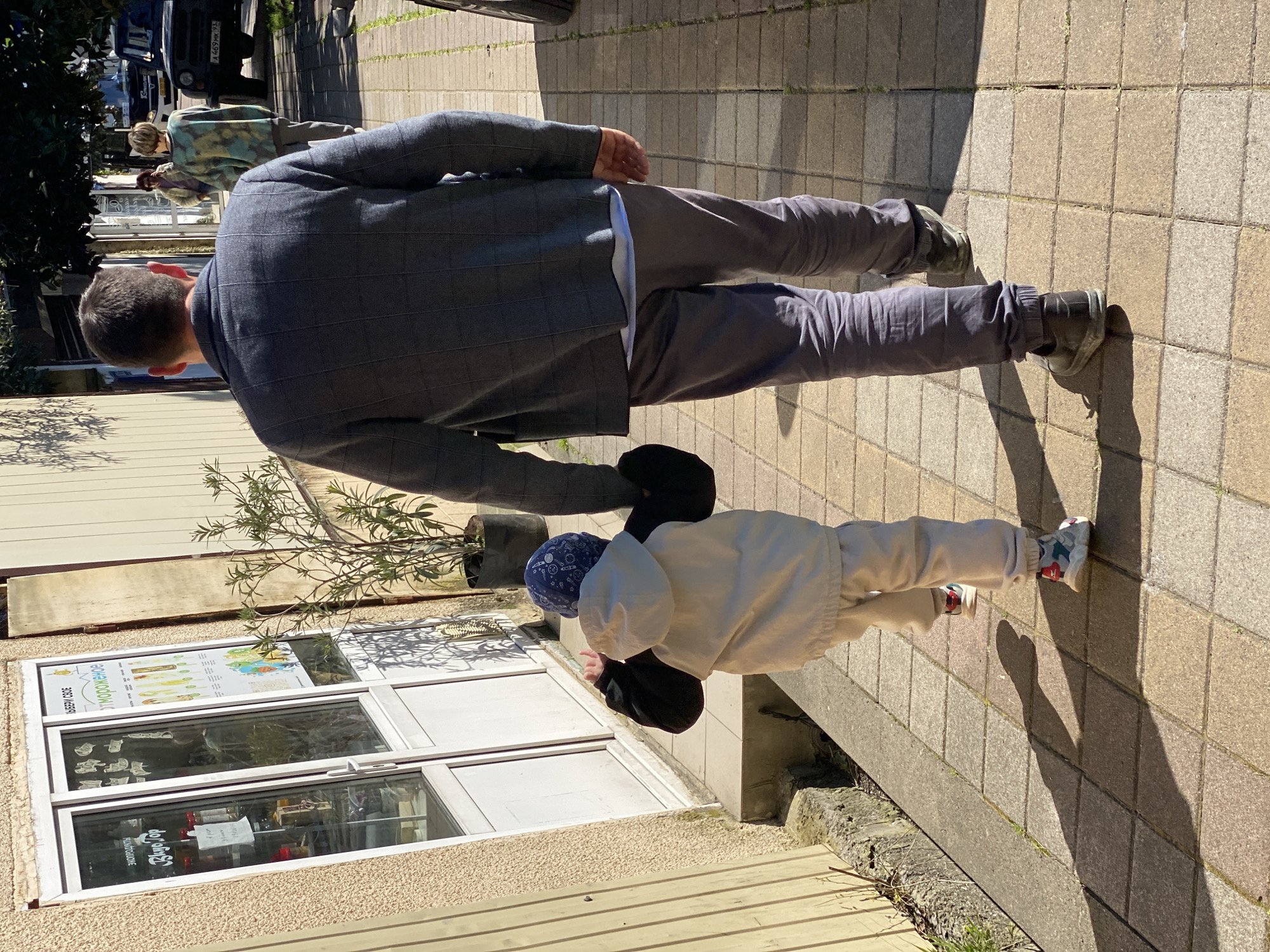 The image depicts a man and a child walking along a paved sidewalk, likely in the city of Sochi, Russia. The man is wearing a dark gray checkered blazer, gray pants, and dark-colored shoes. The child, who appears to be much smaller, is wearing a light-colored jacket and pants and sneakers. The child also has on a blue bandana. Both are walking on a sidewalk paved with tan-colored square tiles.
Behind them is a building with a basement or cellar window visible, with advertisements and possibly product displays inside. Vegetation is visible in a planter and in the background. Other pedestrians and vehicles are faintly visible further in the distance. The shadows indicate it is daytime.