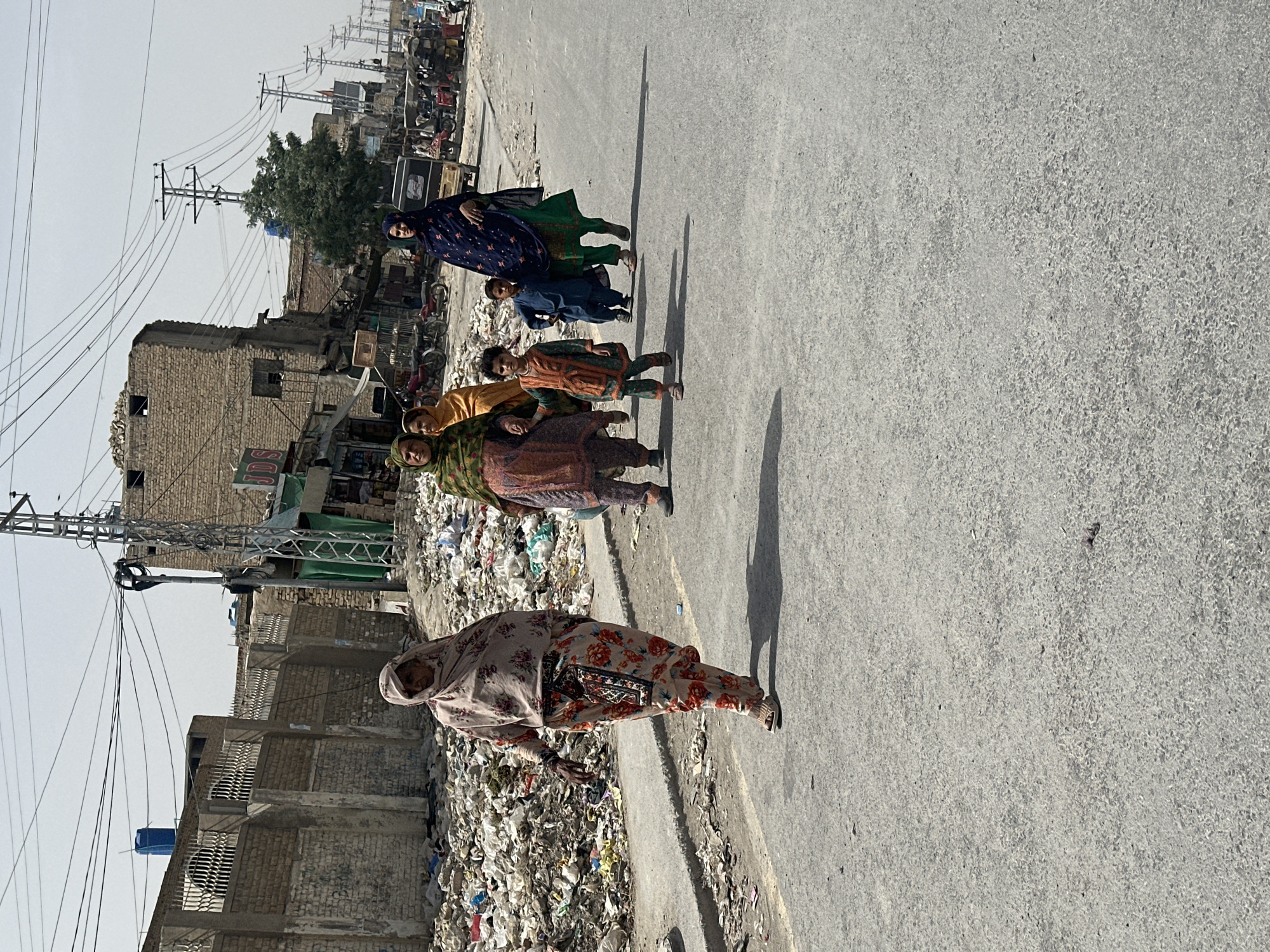 The image depicts a line of women and children walking along a paved street in what appears to be Quetta City Tehsil, Pakistan. The environment is characterized by buildings, utility poles with numerous wires, and a significant amount of refuse and trash lining the edges of the road. The people are dressed in traditional clothing, and their expressions and actions suggest a common purpose. The lighting implies a bright, sunny day.