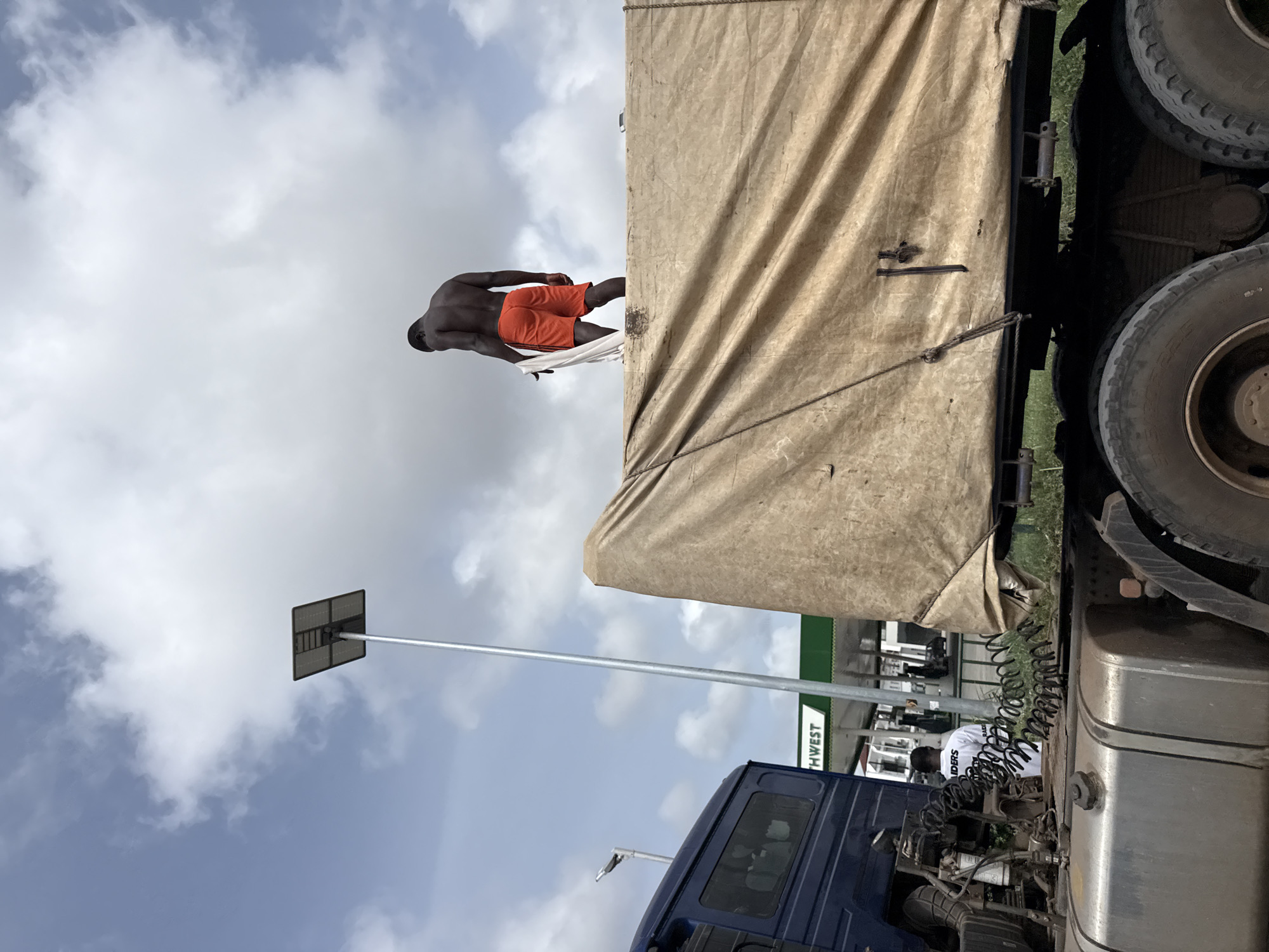 The image depicts a shirtless, dark-skinned man wearing orange shorts standing on top of a large truck with a tan canvas cover. The truck is parked at a gas station in Lagos, Nigeria. Another man in a white shirt is working near the truck's engine. A solar-powered street lamp is visible in the background. The sky is cloudy. The overall composition is from a low angle, looking upwards.