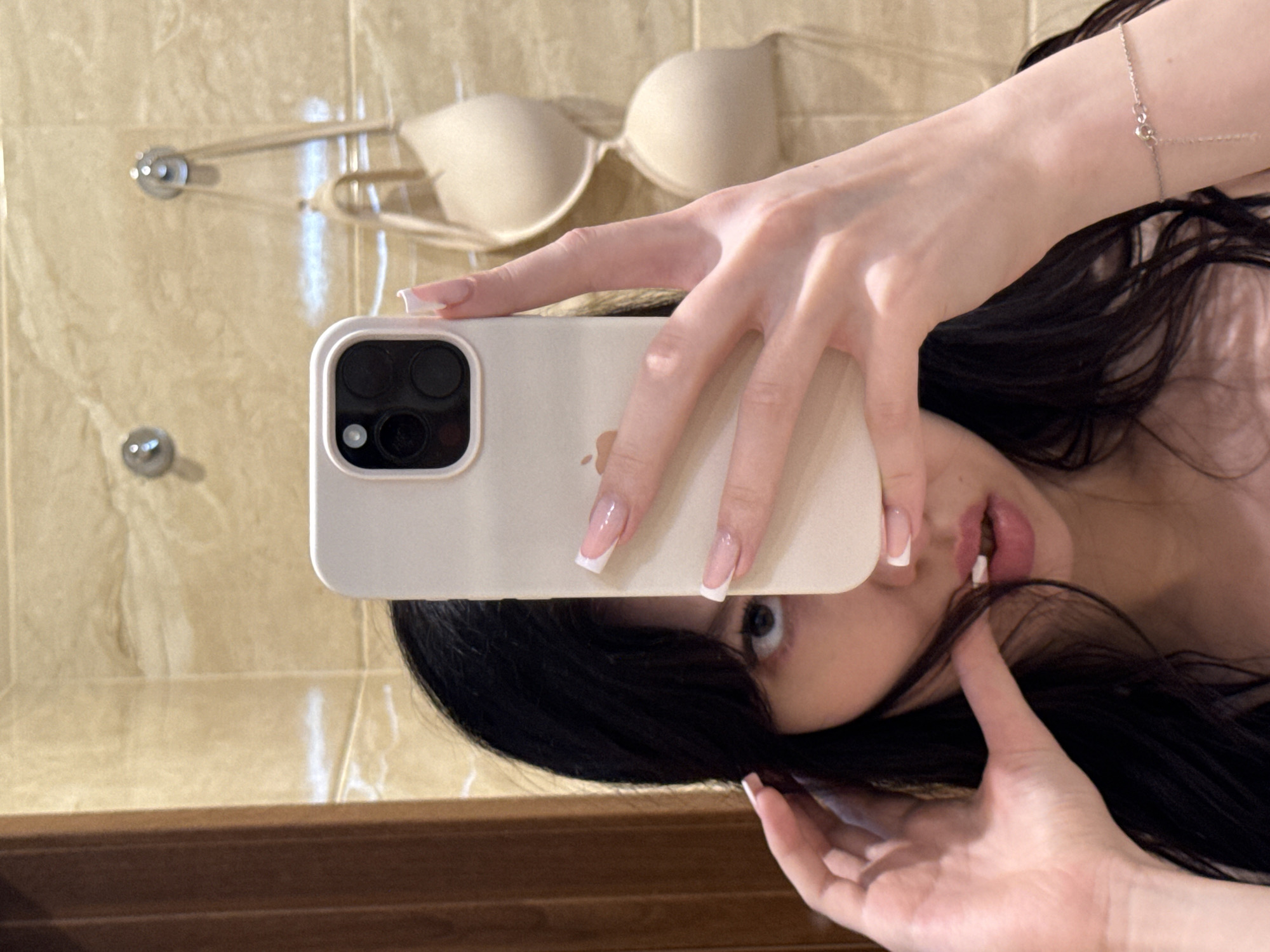 A person is taking a selfie in a bathroom. The person has long dark hair and is holding a phone in their right hand. Their left hand is touching their hair near their mouth. They are wearing a bracelet on their left wrist. A beige bra is hanging on the wall behind them. The wall appears to be tiled with a marble-like pattern. The person's face is partially obscured by the phone. The environment suggests a residential bathroom, possibly in an apartment in Khimki, Russia, given the provided location context.