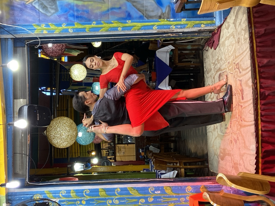 A man and woman are engaged in a tango dance within a colorful setting. The woman wears a vibrant red dress, while the man is dressed in a dark vest and trousers. They are surrounded by hanging lights and a backdrop of brightly painted surfaces, including blue and yellow geometric patterns. The scene is indoors, possibly a restaurant, with tables and chairs visible in the background.