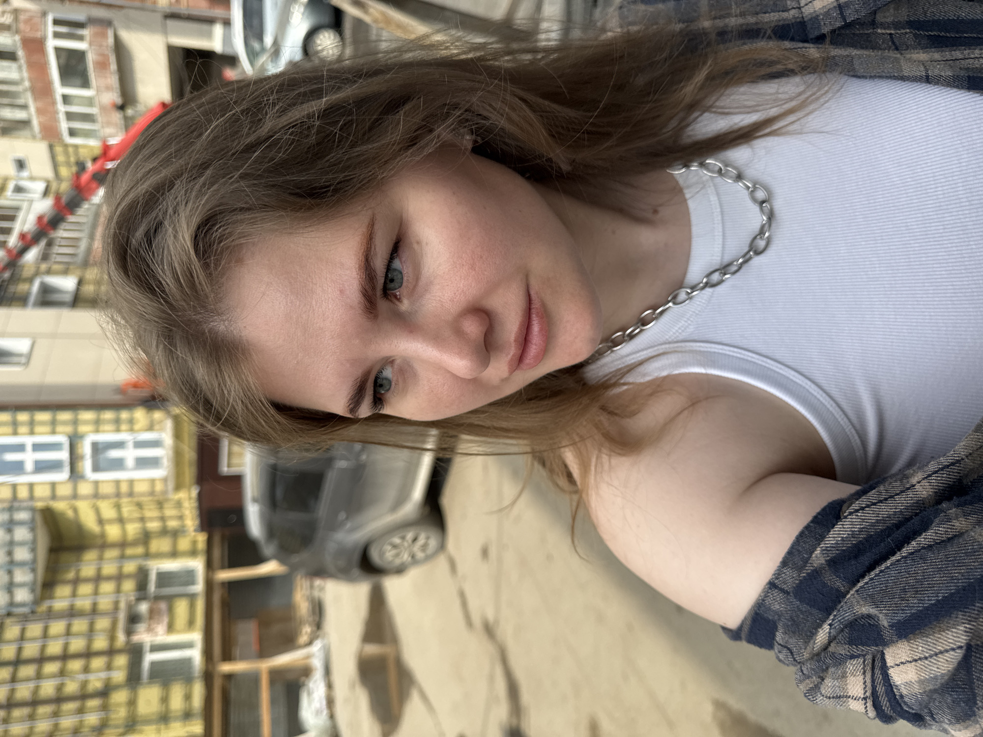 A young woman with light brown hair is centered in the image, wearing a white tank top and a silver chain necklace, with a plaid shirt visible on her right arm. The background shows a street scene in Perm, Russia, with multi-story buildings, including one under construction with yellow insulation panels, and a parked gray car. A red construction crane is also visible behind the buildings.