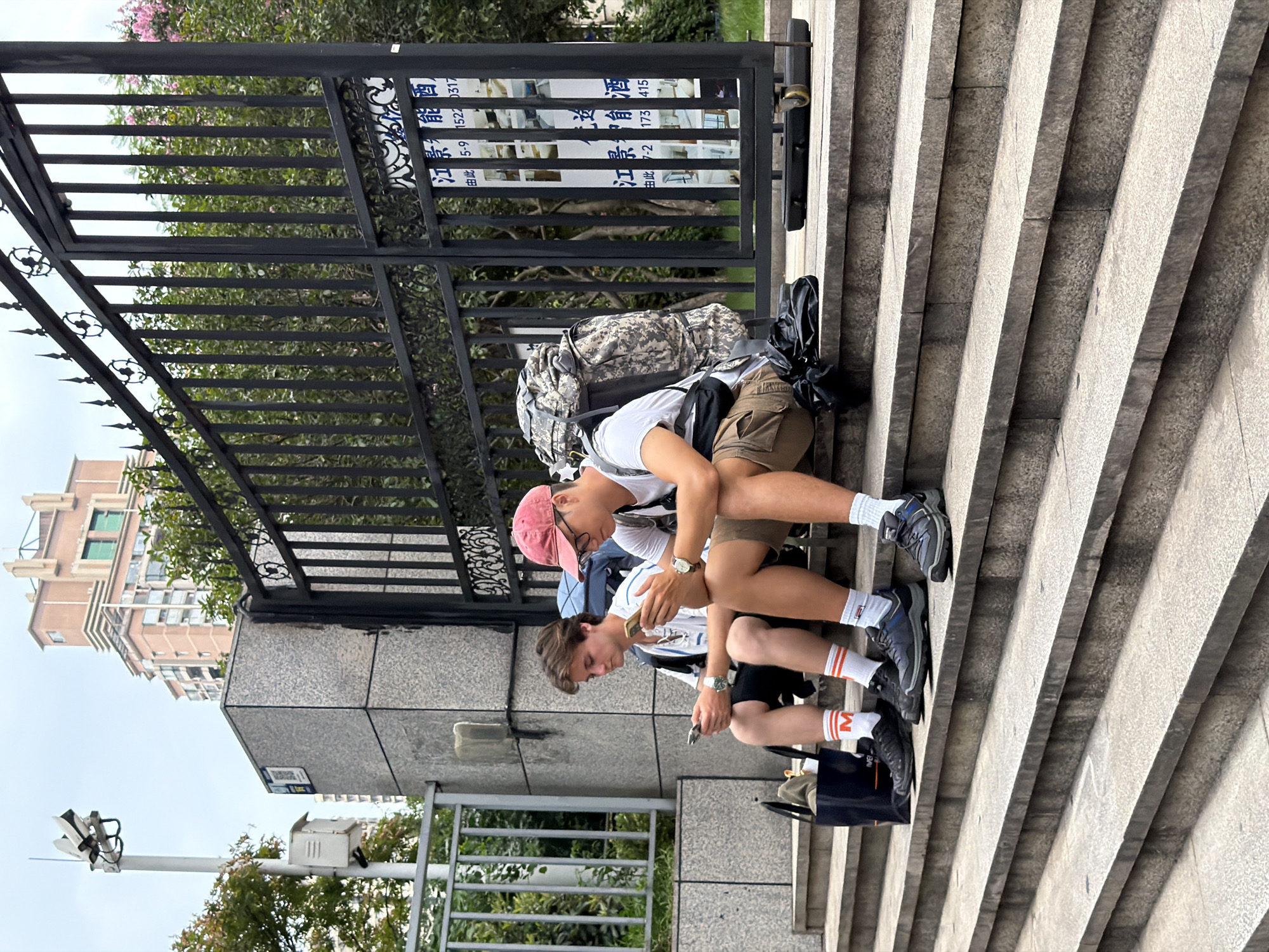 Two young men are sitting on stone steps outdoors, facing each other and looking at their phones. The man on the left has light brown hair and is wearing a white t-shirt with black trim, black shorts, and white socks with orange stripes. He has a black bag with a beige item on top next to him. The man on the right has darker brown hair and is wearing a red baseball cap, glasses, a white t-shirt, khaki shorts, and white socks. He has a large, camouflaged backpack with him. Behind them is a black metal gate with decorative scrollwork and several signs with Chinese writing. In the background, there are trees and a multi-story building. The scene appears to be in the Yuzhong District of China.