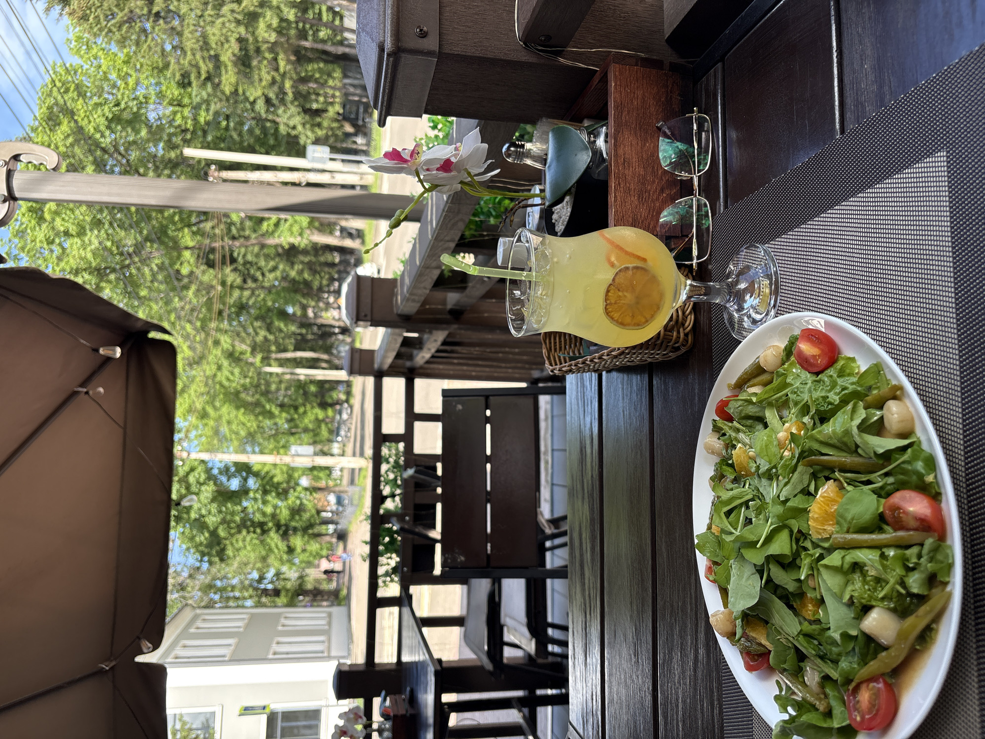 A sunlit outdoor seating area of a restaurant in Birobidzhan, Russia, features a table set for a meal. On the table, a white oval plate holds a vibrant salad composed of green leafy vegetables, cherry tomatoes, asparagus spears, orange segments, and what appear to be small white cheese cubes. Beside the salad is a tall stemmed glass filled with a yellow beverage, garnished with a slice of orange and a green straw. Also on the table are a pair of glasses with gold frames. In the background, a wooden railing encloses the seating area, beyond which trees and a building are visible, suggesting a city setting. The sky is blue with some clouds. The overall scene depicts a relaxed outdoor dining experience.