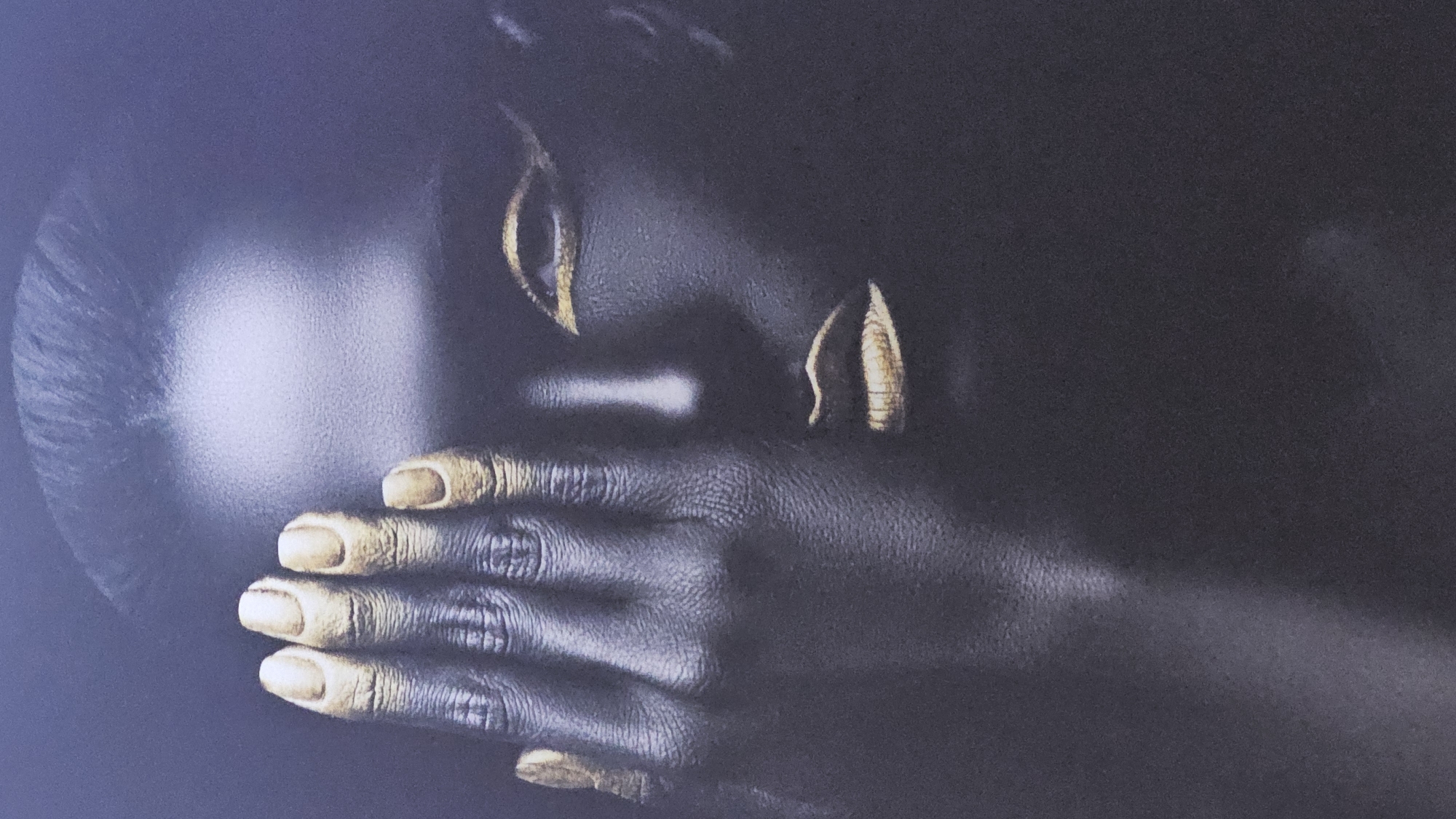 The image is a close-up of a person with dark skin. The person's hand is covering their mouth. The fingernails and earrings are painted gold. The background is dark and indistinct. The location is not evident in the image.