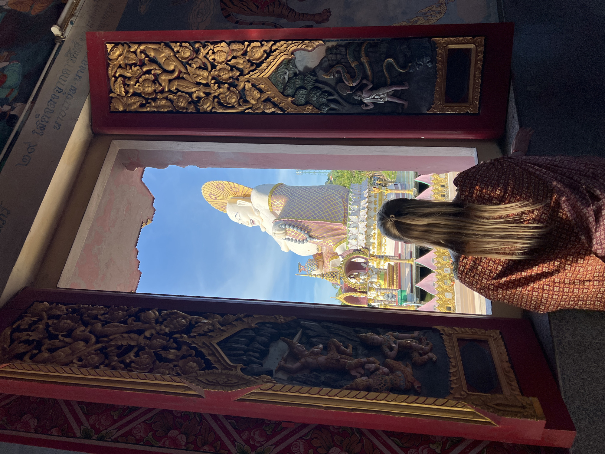 Captured in Baan Plai Laem, Thailand, this photograph reveals a scene viewed through an ornate window. Intricately carved wooden panels frame the view of a large, white Buddha statue under a blue sky. A woman with long, blonde hair, clad in a patterned orange top, stands near the window, partially obscuring the view of the statue. The interior space around the window exhibits elaborate Thai-style decorations.