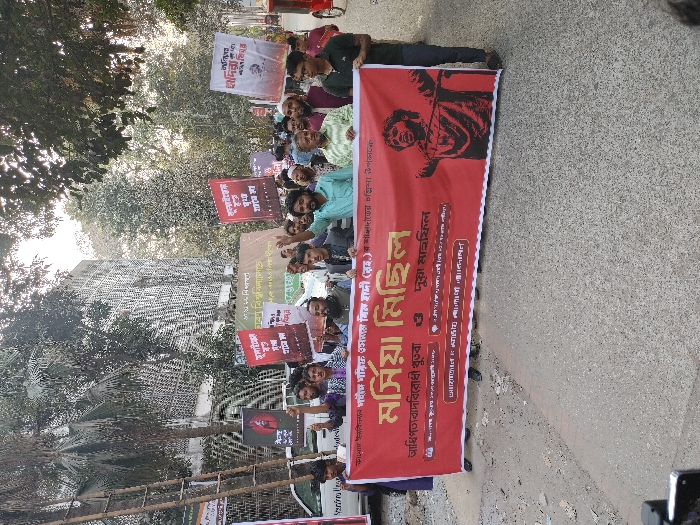 This image captures a daytime protest or rally on an urban street in Dhaka, Bangladesh.
Approximately 20-30 individuals, primarily adult males, are depicted marching or standing in a procession. At the forefront, several participants hold a large, horizontal red banner, approximately 10-12 feet in width. This banner displays prominent white Bengali text reading "মর্সিয়া মিছিল" (Mourning Procession/Elegy Rally) and includes smaller Bengali text referencing "Anti-Hegemony Speech" and "Prayer Meeting". On the right side of this main banner, a stylized black and red graphic depicts a man with outstretched arms.
Behind the main banner, numerous participants carry smaller, vertical rectangular banners and posters. Many of these are red with white Bengali text; one is white with black text. Some feature graphic elements, such as a silhouetted face on a red background. Several individuals in the procession are shown with their right fists raised in the air. Participants are dressed in a mix of casual shirts, t-shirts, and traditional kurtas.
The street surface is paved, and a raised curb or sidewalk is visible on the left. In the background, dense green foliage from trees lines the street, partially obscuring urban buildings. One visible building features a distinctive, light-colored geometric patterned facade. A white vehicle is partially visible on the far left, and a red auto-rickshaw (CNG) is partially visible on the far right. The scene is illuminated by natural daylight.