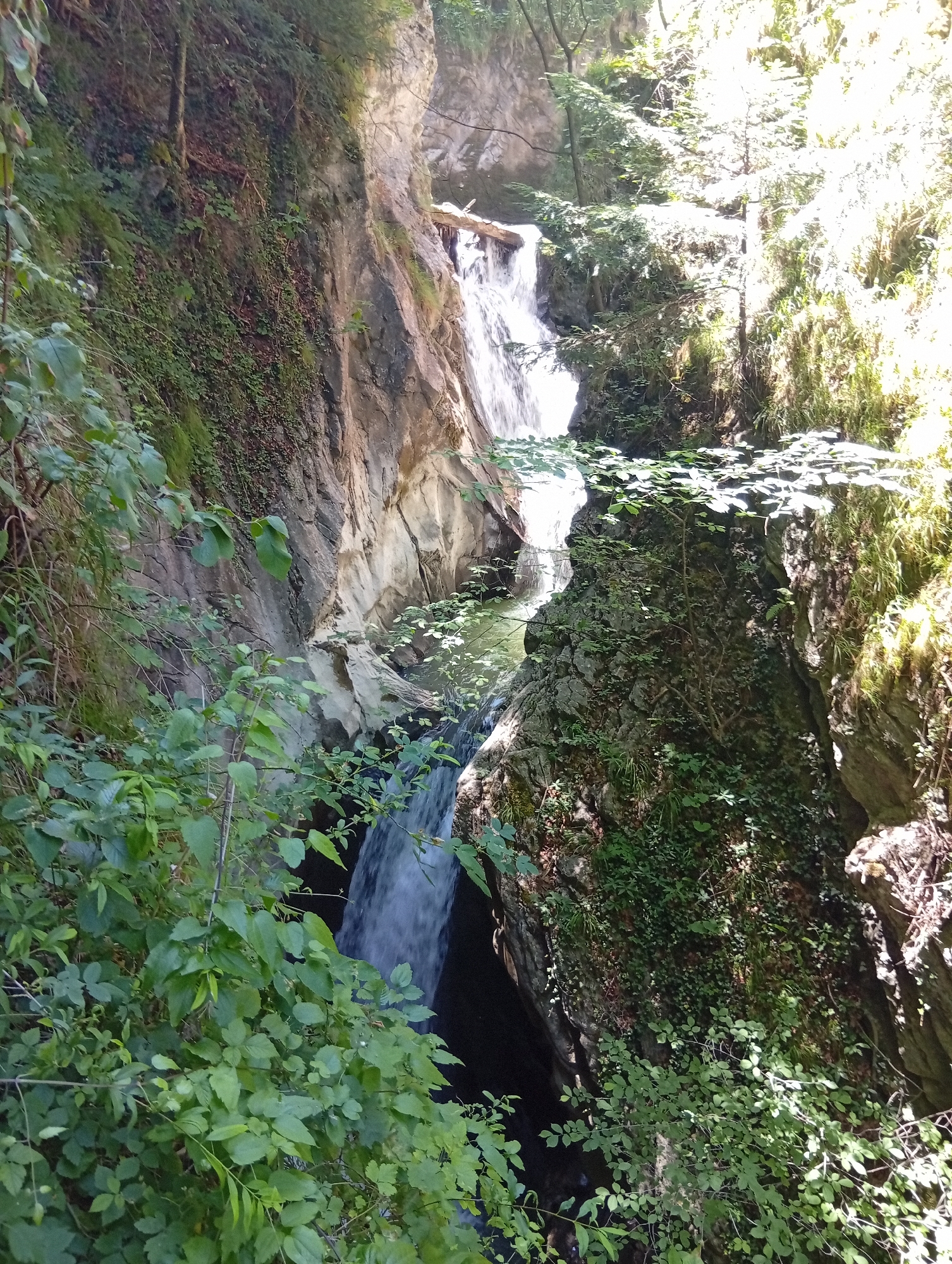 The image depicts a natural scene with a waterfall cascading down rocky terrain. Lush green vegetation covers the surrounding cliffs and slopes. Sunlight filters through the trees, illuminating parts of the scene. The waterfall appears to be a focal point, with water flowing into a pool or stream below. The overall environment is rugged and natural, suggesting a remote or undeveloped area. No people or man-made structures are visible.