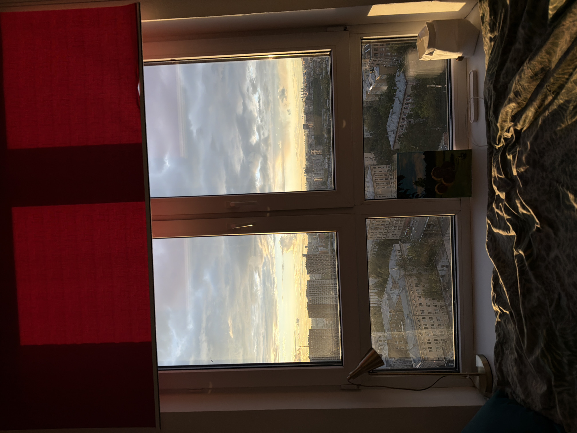 Here is a description of the image:
The image captures an interior view of a window looking out onto a city at what appears to be dawn or dusk. The window is divided into four panes, with the left side of the window partially covered by a red curtain or blind. The view through the left two panes reveals a sky filled with clouds, catching the colors of the rising or setting sun, illuminating the city skyline in the distance. The buildings are silhouetted against the bright sky. The right two panes of the window offer a closer view of the city, showing buildings and trees. On the windowsill, there's a lamp and what appears to be a small painting or framed picture. A curtain or fabric is draped on the right side of the window, partly obscuring the view.
