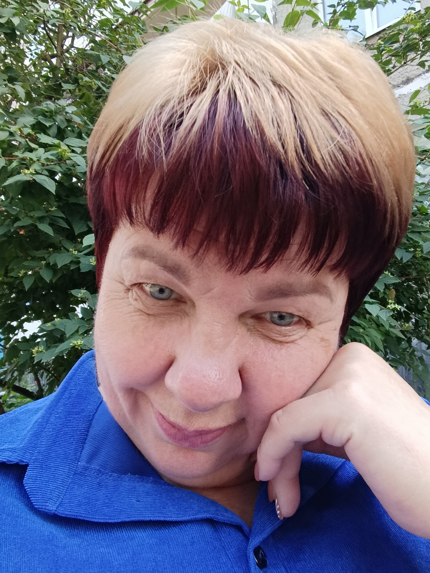 The image is a close-up, selfie-style portrait of a middle-aged woman with blonde and dark red hair. She is wearing a blue collared shirt. Her right hand is resting on her chin, and she is looking slightly down and to her left with a gentle smile. The background consists of green foliage, suggesting an outdoor setting, likely a garden or park, possibly in Svietlahorsk, Belarus. The lighting appears to be natural daylight.