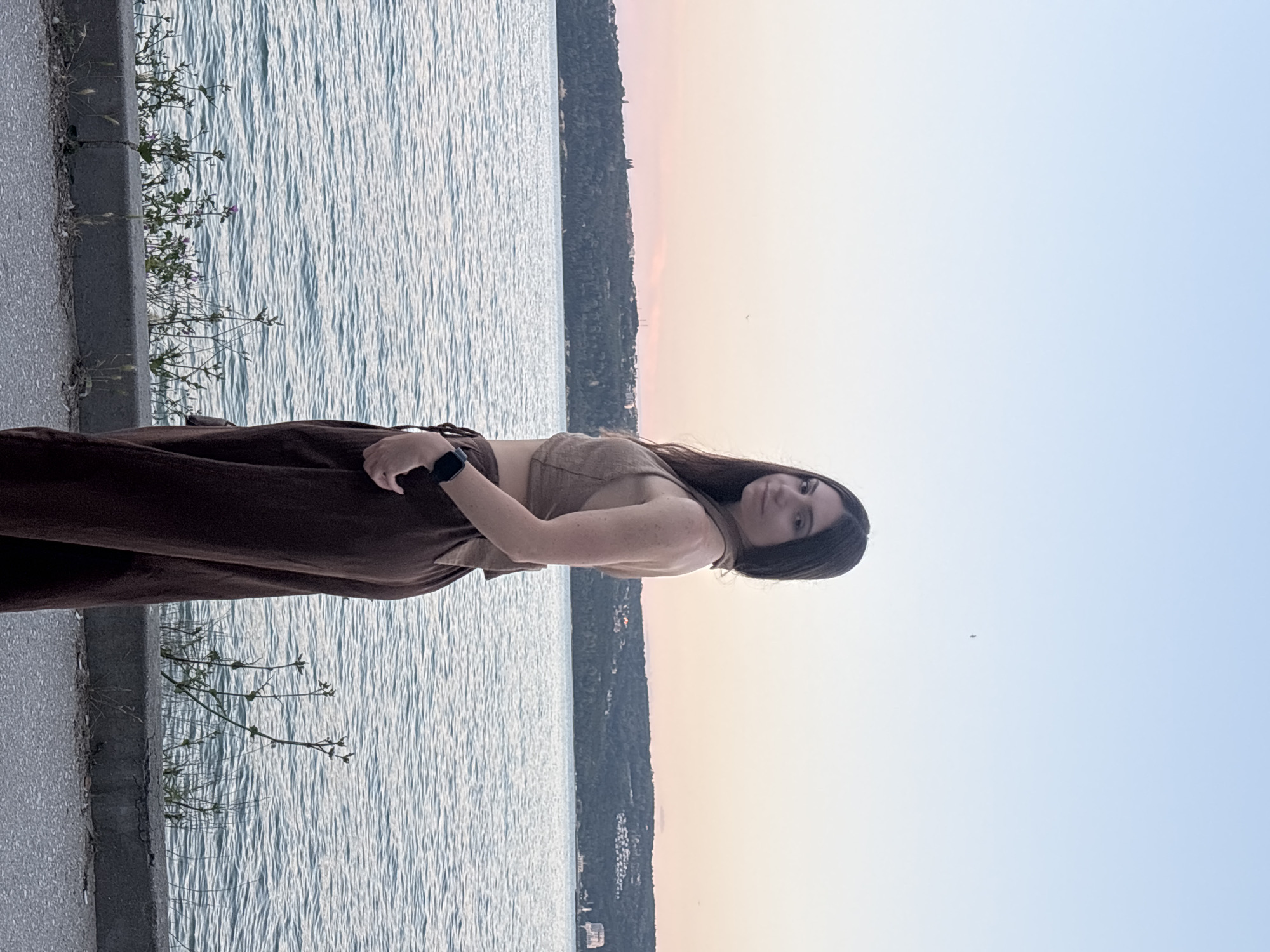 A young woman with long dark hair stands on a paved walkway next to a body of water. She is wearing a brown halter top and brown wide-leg pants. The water behind her is rippled, and a green, treed hill with buildings is visible in the distance. The sky above is a gradient of light blue and soft pink, indicating it is either sunrise or sunset. The scene appears to be on the Varna Sea Garden promenade, a popular waterfront park in Varna, Bulgaria.
