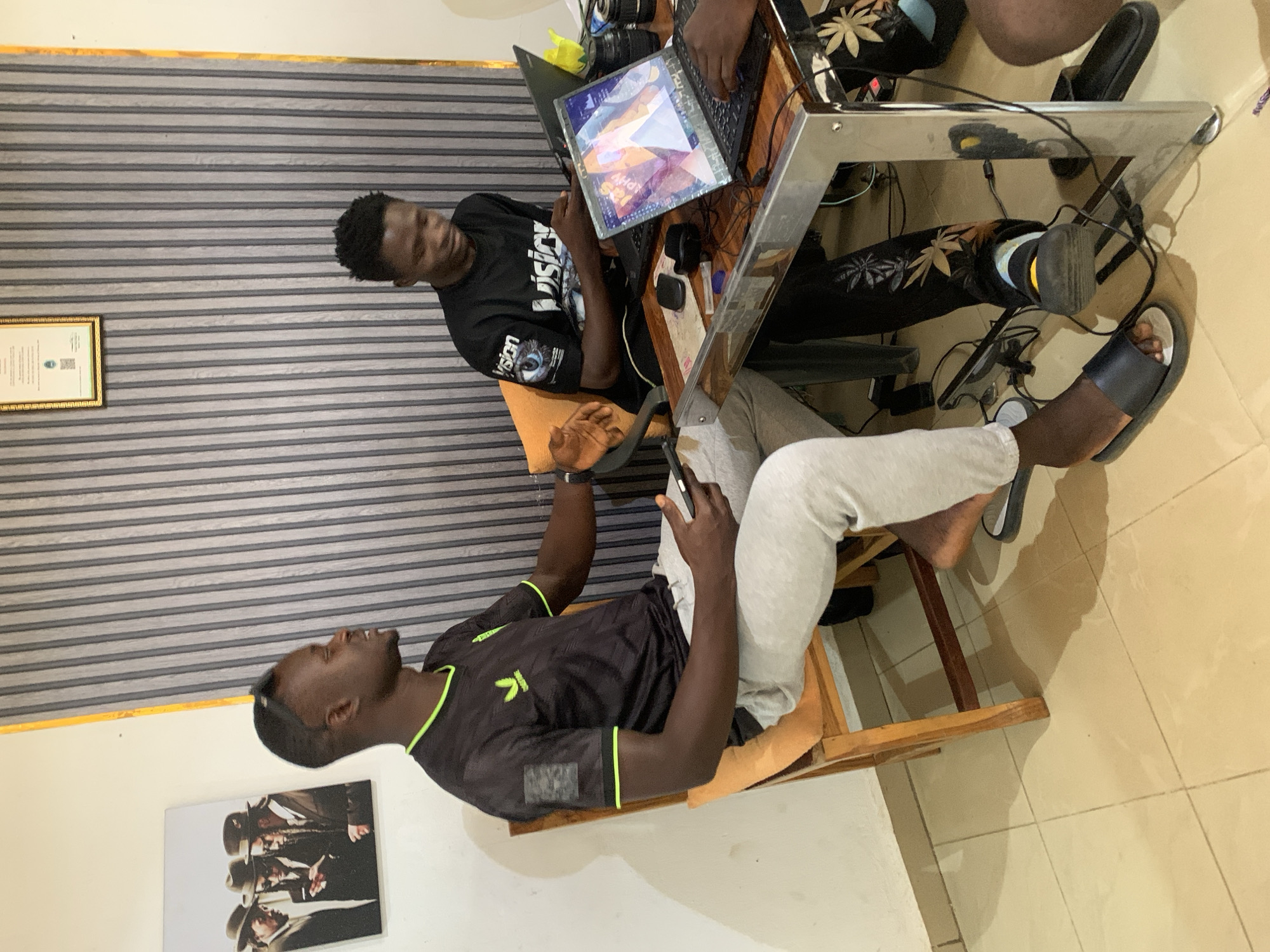 Three African men are in an indoor space, possibly a home or office, likely located in Jos, Nigeria. One man, wearing a black t-shirt with a "Vision" print, black pants with leaf designs, and yellow sandals, is seated, facing away from the camera. Another man, wearing a black t-shirt with green stripes and grey sweatpants, is sitting on a chair and looking upward. The third man is partially visible, also seated. All three are gathered around a metal-framed table with two laptops and other items such as a mouse, camera lenses, and cords on it. The room features striped wallpaper, a framed certificate on the wall, and a framed poster with a black and white photo. The floor is tiled in cream.