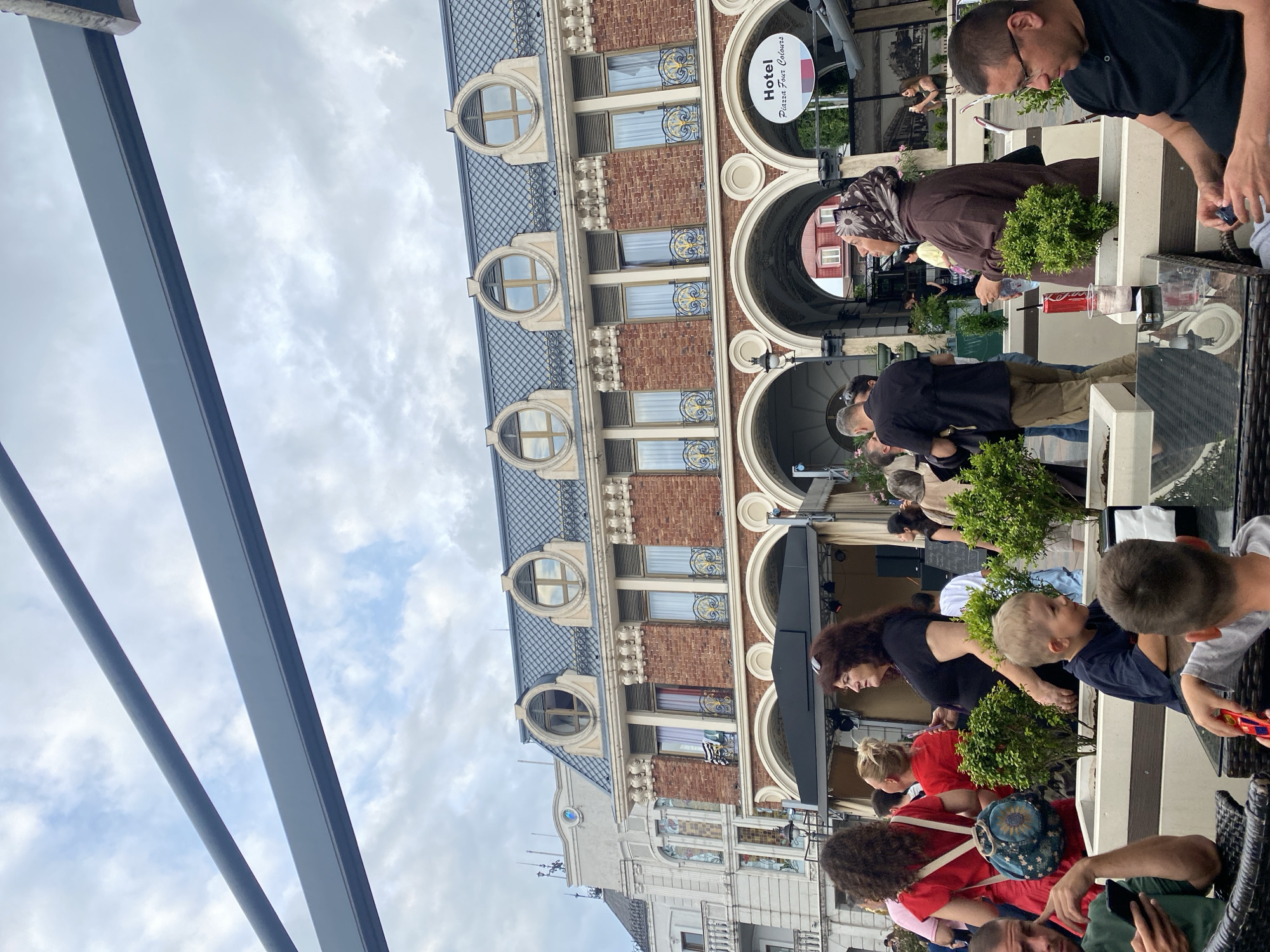This media file captures an outdoor scene at a cafe in Batumi, Georgia. A building with arched entrances and ornate circular windows forms the backdrop. The building's facade is a mix of brick and lighter stone, with a dark tiled roof. Above one of the arches, a circular sign reads "Hotel Piazza Four Colours".
Several people are seated at tables, some appearing to be enjoying drinks. In the foreground, a young boy with blond hair wearing a dark blue shirt is looking to the right, seemingly engaged with something on a table. To his left, a woman with long dark hair and a red shirt is also present. Further back, other individuals are visible, including a woman wearing a headscarf and a brown garment, and a man with glasses in a black shirt. The atmosphere suggests a casual daytime gathering. Lush green bushes are placed around the seating area, adding to the outdoor ambiance. The sky is cloudy, indicating overcast weather. The overall scene depicts people relaxing at a cafe in an urban European setting.
