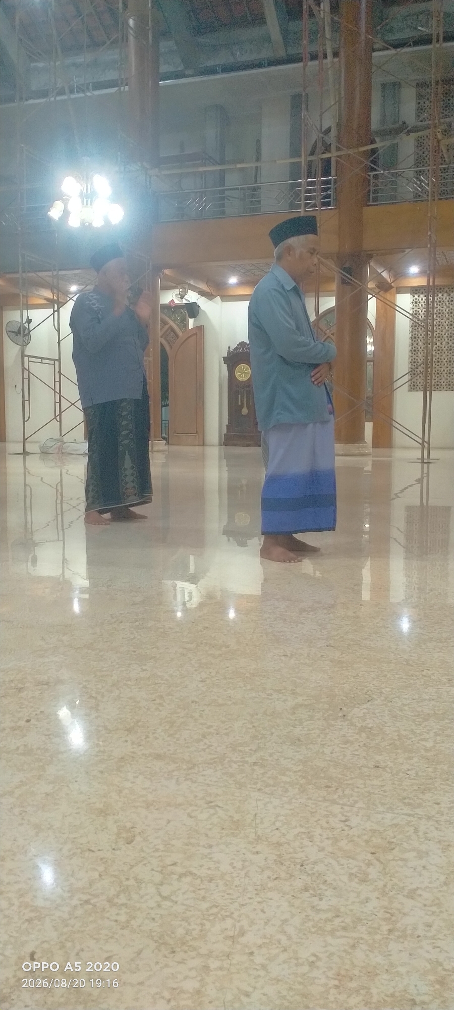 In a large, indoor space with a high ceiling, two men are standing on a polished, reflective floor. The man on the left is wearing a dark patterned sarong and a blue long-sleeved shirt, with a black peci (cap) on his head. He is facing to his left, with his hands clasped in front of him. The man on the right is wearing a light blue long-sleeved shirt and a blue and white sarong, also with a black peci. He is facing to his right, with his arms crossed.
The environment appears to be a building under construction or renovation, with scaffolding visible in the background. A large chandelier hangs from the ceiling on the left. To the right of the second man, a grandfather clock with an ornate wooden case is visible. Doors and patterned screens are also present in the background. The floor reflects the lights from the chandelier and other sources, creating bright spots. The timestamp on the bottom left corner indicates the photo was taken on August 20, 2026, at 7:16 PM, and was captured by an "OPPO A5 2020" device. The location is identified as Kedung, Indonesia.
