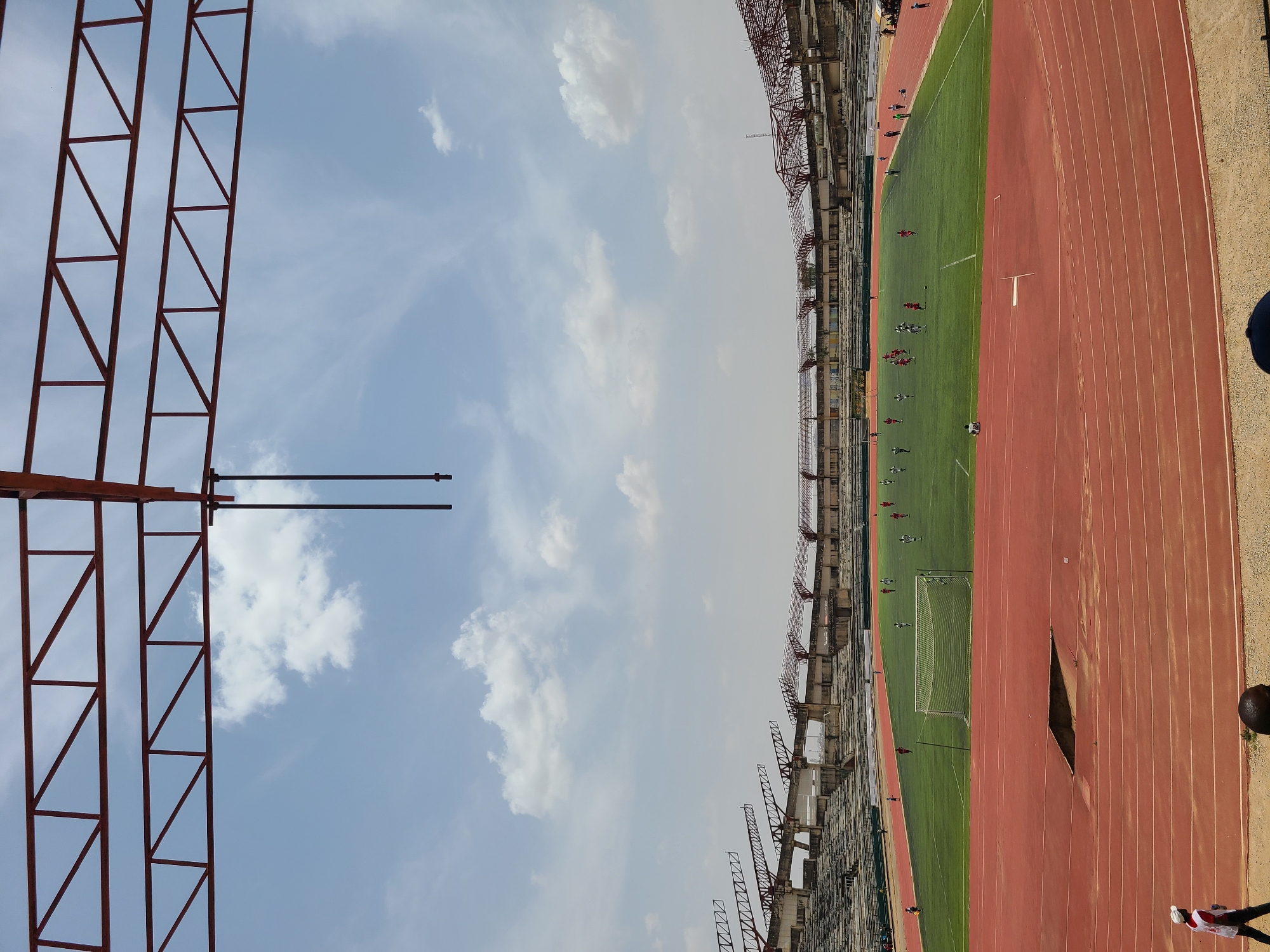 An outdoor sports stadium is depicted from an elevated perspective, oriented with the blue sky and scattered white clouds occupying the upper and left portions of the frame. Dominating the left foreground are large red metallic truss structures, likely part of the stadium's framework, such as for lighting or a roof support system.
In the midground and background, the extensive concrete structures of the stadium's seating areas are visible, appearing somewhat aged or in a state of construction/renovation. The right side of the image features a vibrant green football (soccer) pitch, where several individuals, presumably players, are actively engaged in a game or practice session. Some players are distinguishable by their red jerseys, while others wear lighter colors. A white goalpost is clearly visible on the pitch.
Encircling the green field is a red athletics running track, complete with distinct white lane markings. Notably, there is a triangular dark opening or damaged section on the track in the mid-right. Beyond the track to the far right, a sandy or dirt area is visible. A person wearing a white shirt with red accents and dark pants is partially visible at the bottom right corner, suggesting spectators or staff. Another person is visible further right, above the track.
The scene takes place during daytime, under bright, clear skies with minimal shadows, indicating the sun is high. The overall setting is a large sports complex, consistent with the provided location context of a "New Stadium, Nigeria." No discernible text is visible in the image.