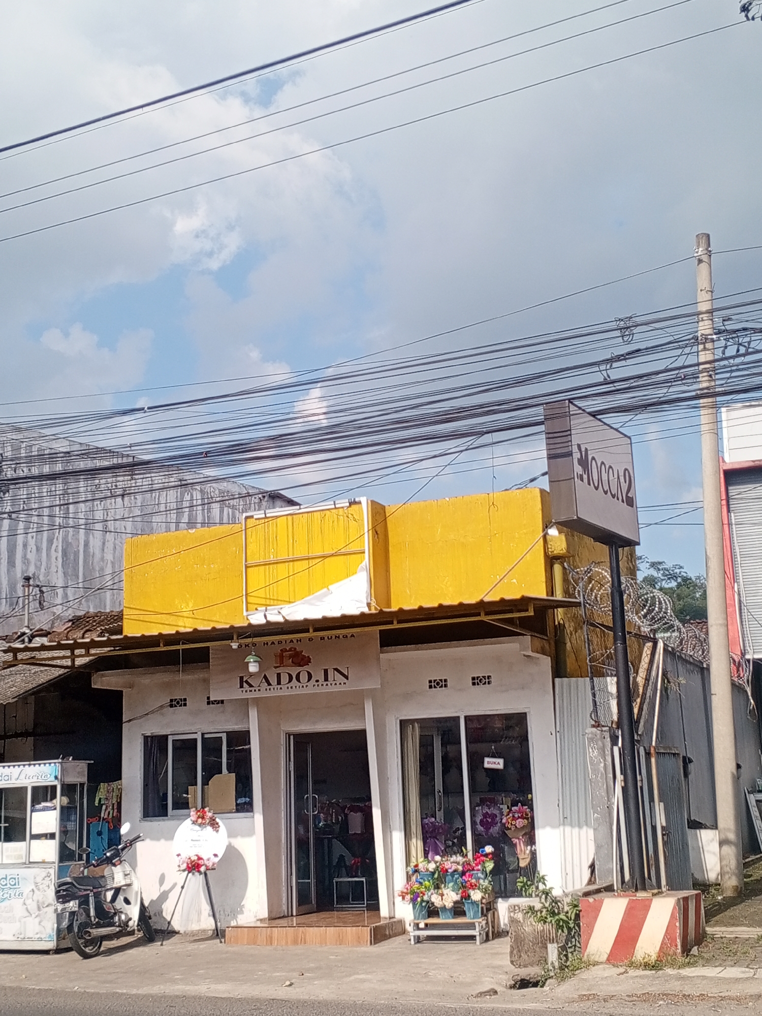 This media file depicts a storefront in Sumbersari, Indonesia. The building is painted white with a bright yellow awning above. A sign in front of the store reads "KADO.IN" and below it, "TEMAN SETIA SETIAP PERA", which translates to "Trusted Friend for Every Celebration". Above the awning, another sign displays "MOCCA2". The storefront has large glass windows and a doorway, with flower displays both inside and outside the shop. To the left of the store, a food cart or stall is visible, with text that appears to say "dai Lteerta" and "doi rla". A motorcycle is parked in front of the building. The scene is set outdoors, under a bright sky with scattered clouds, suggesting it is daytime. Overhead, numerous electrical wires are strung across the frame, a common sight in many urban and semi-urban areas. Barbed wire fencing is visible behind the store. The overall impression is of a small business operating on a street.