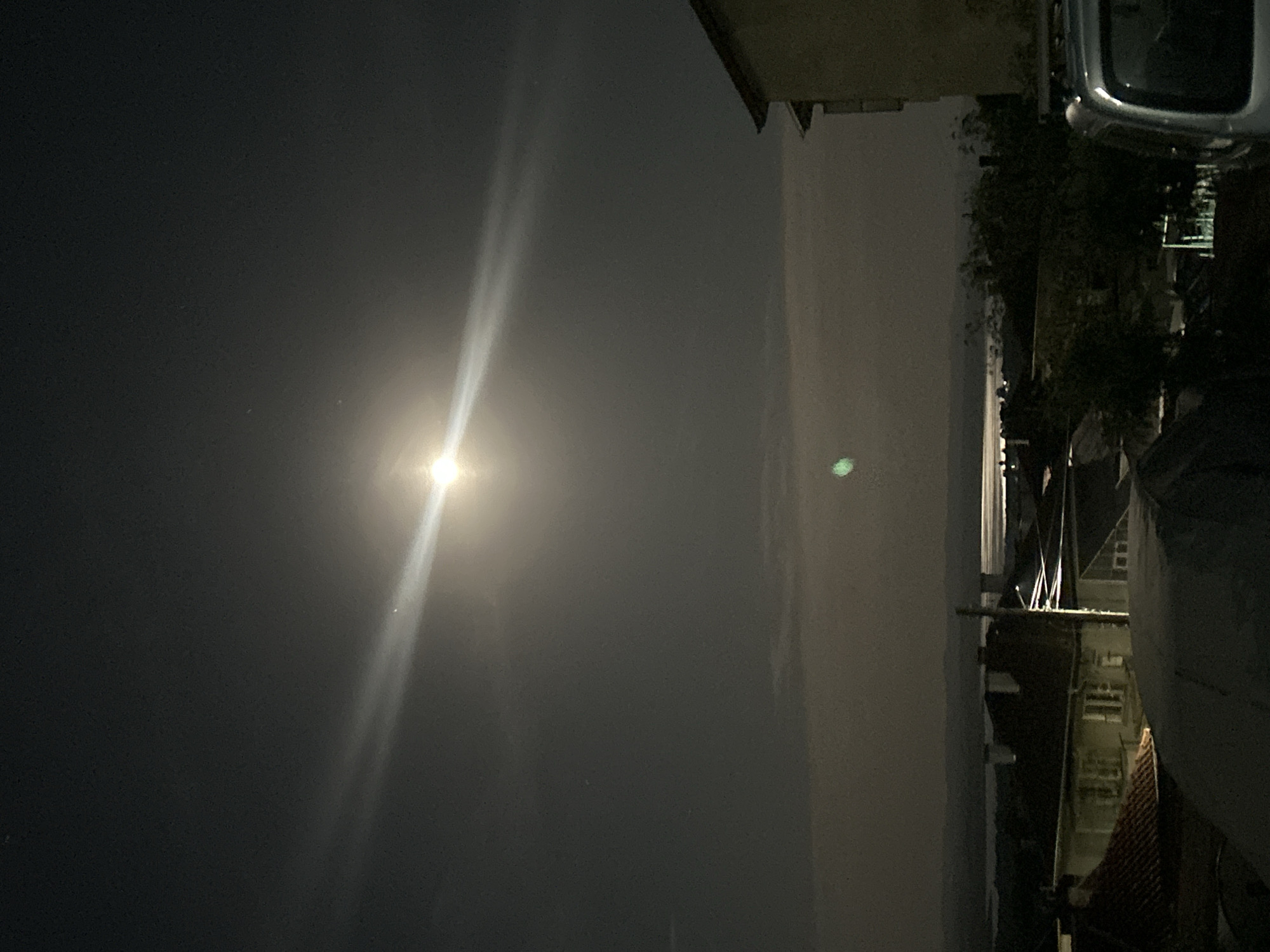 A night scene. A bright light is visible in the upper left quadrant, possibly a street lamp or the moon, with beams extending from it. The right side of the image shows a residential area, with buildings lit by streetlights. A silver car is parked near the buildings. The scene appears to be captured from an elevated position, possibly a rooftop or upper floor. Without any other identifiers, pinpointing the exact location in Burgas, Bulgaria is not possible.