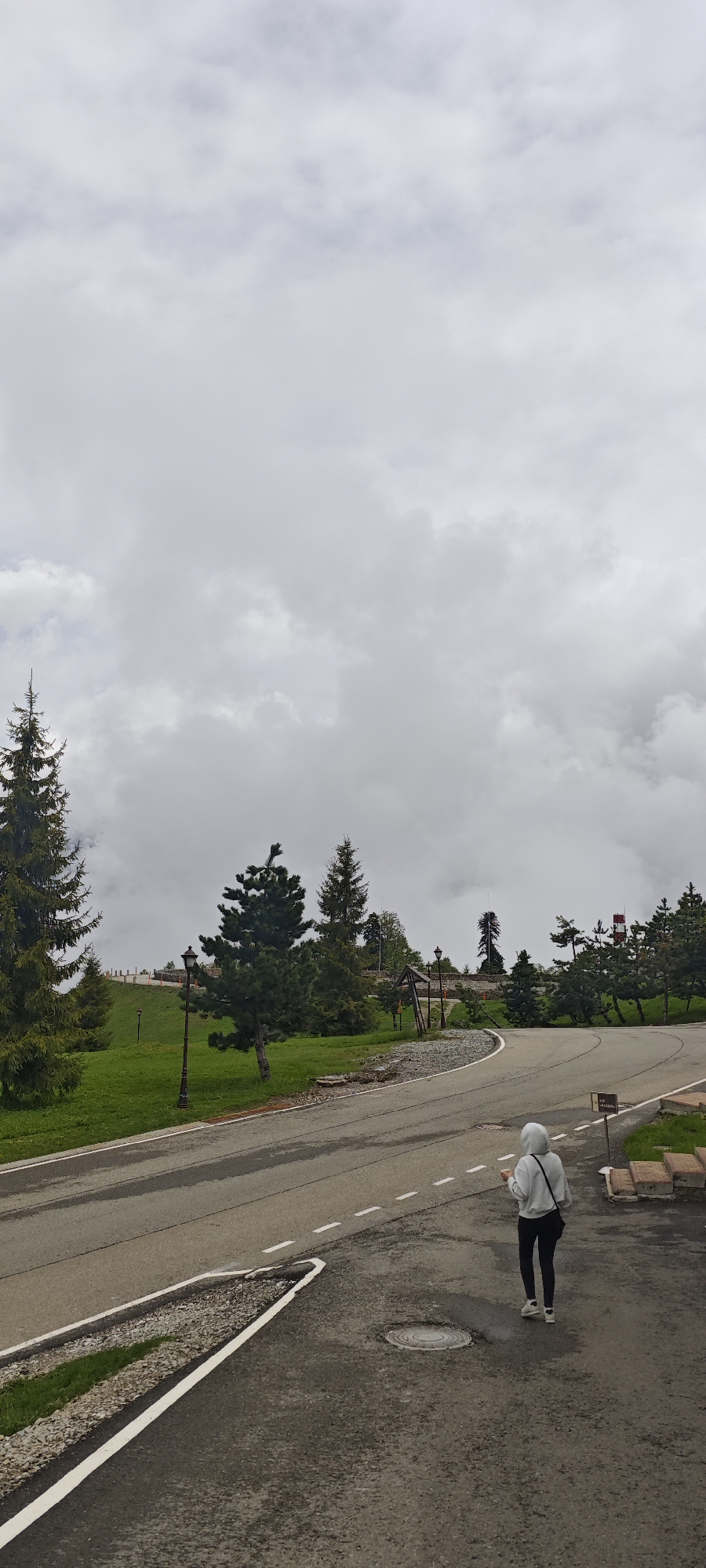 A long shot shows a person walking on an asphalt road next to a green grassy area and trees beneath a cloudy sky.
The road, made of gray asphalt, curves slightly to the right. A white dashed line marks the center. A person in a gray hoodie, black pants, and white sneakers walks away from the viewer along the road's right side.
The left side of the road features a grassy area dotted with trees of varying heights. A black lamppost stands near the roadside. Additional trees and a wooden structure are visible further up the slope. A white-and-red tower can be seen in the distance, partially obscured by trees.
The sky is overcast with heavy gray clouds. The scene is situated in an unknown location in Russia.