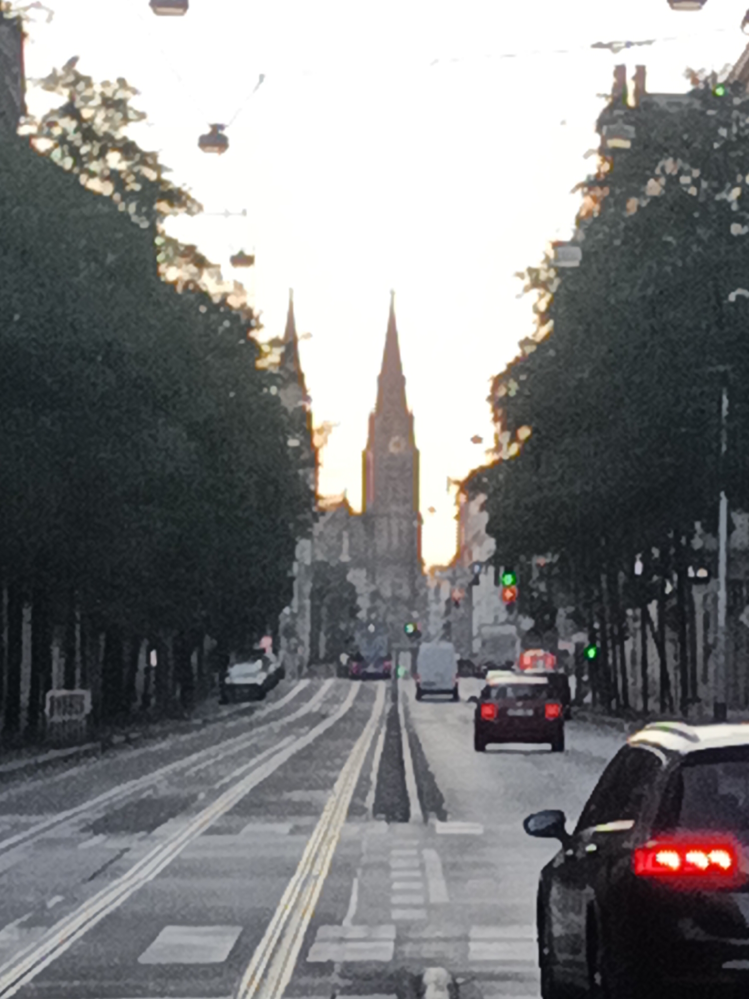 The image captures a street scene in Prague, Czechia, with a prominent church spire in the background, likely part of the Church of St. Nicholas in Old Town Square or another significant religious building with a similar architectural style. The perspective is from a street with tram tracks running down the center. On either side of the street, there are tall, mature trees, creating a canopy effect. The sky is bright, suggesting either early morning or late afternoon, with the sun's rays filtering through the trees and illuminating the scene.
Several vehicles are visible on the road. In the foreground, a dark-colored car is seen from the rear, its brake lights illuminated. Further down the road, other cars, including a red hatchback and a white van, are also present. Traffic lights with green signals are visible. The road surface is paved, and the tram tracks are clearly defined. Pedestrians are not distinctly visible, though a blurred figure can be seen near the left side of the road. The overall atmosphere is that of a city street during daylight hours, with elements of historical architecture and urban infrastructure.