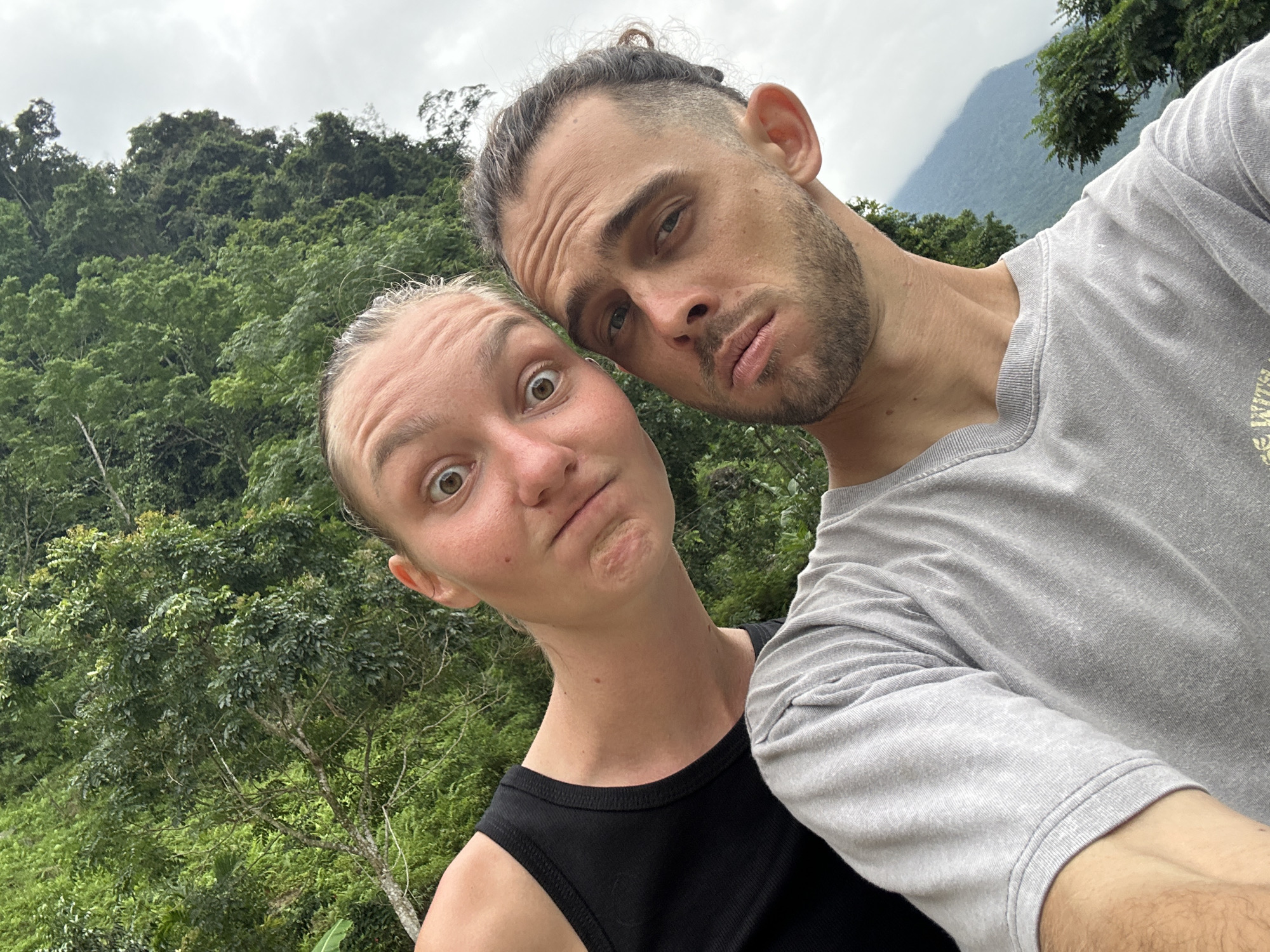 In a selfie taken outdoors, two individuals are prominently featured. The person on the left, a woman, is leaning towards the camera with wide, surprised eyes and a pursed mouth. She is wearing a black tank top. To her right, a man is looking directly at the camera with a slightly pensive expression. He has dark hair pulled back into a bun, a beard, and is wearing a grey t-shirt. The background consists of lush green trees and foliage, characteristic of a mountainous or forested area. In the distance, a misty mountain range is visible under an overcast sky. The image appears to be taken in Pù Luông Commune, Vietnam, given the lush, mountainous landscape.