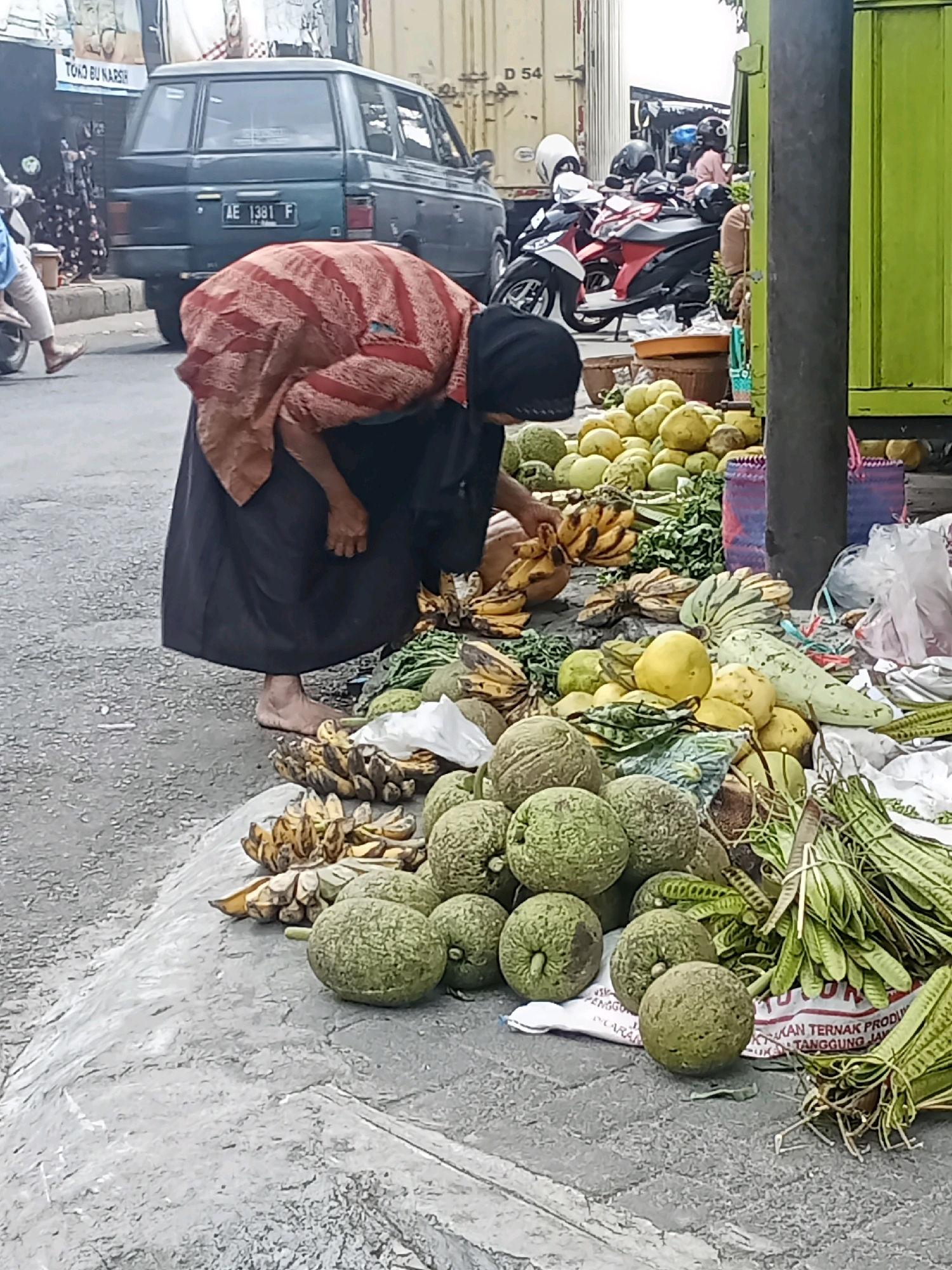 In Sukowinangun, Indonesia, a vendor dressed in a black abaya and a red and white striped headscarf is seen bent over a display of produce on a gray, textured sidewalk. She is barefoot and appears to be arranging or examining the items. The foreground is dominated by a large pile of greenish-gray, rough-skinned fruits, some round and others oblong. To the left, a bunch of ripe bananas with brown spots is visible. Behind the vendor, more fruits, including large yellow melons, are displayed. In the background, a dark-colored SUV with the license plate "AE 1381 F" is parked. Several motorcycles are also present. The scene suggests a bustling outdoor market environment, with a street and buildings visible in the distance, including a tall green structure on the right side of the frame.