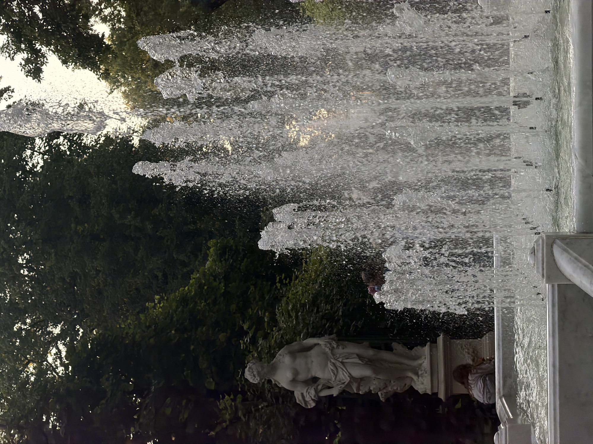 The image shows a fountain in front of a classical sculpture in a park in Saint Petersburg, Russia. The fountain is spraying water high into the air, creating a shimmering effect. The sculpture is of a woman, and it is made of white marble. In the background, there are trees and a building. A young child is sitting on the edge of the fountain's basin, looking at the water. The scene appears to be a tranquil and beautiful day in a park.